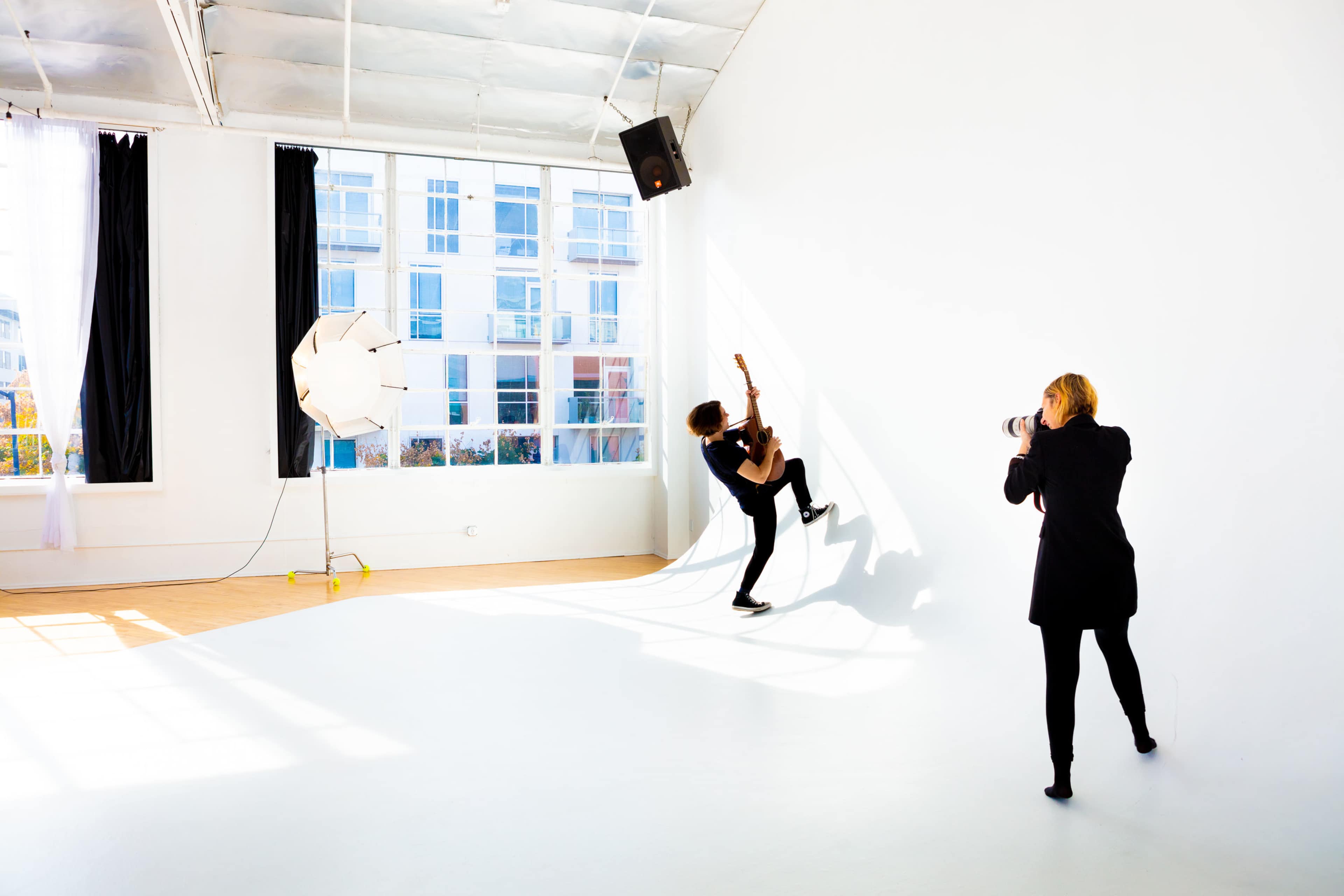 A person plays guitar against a white wall while another person photographs them in a brightly lit studio.