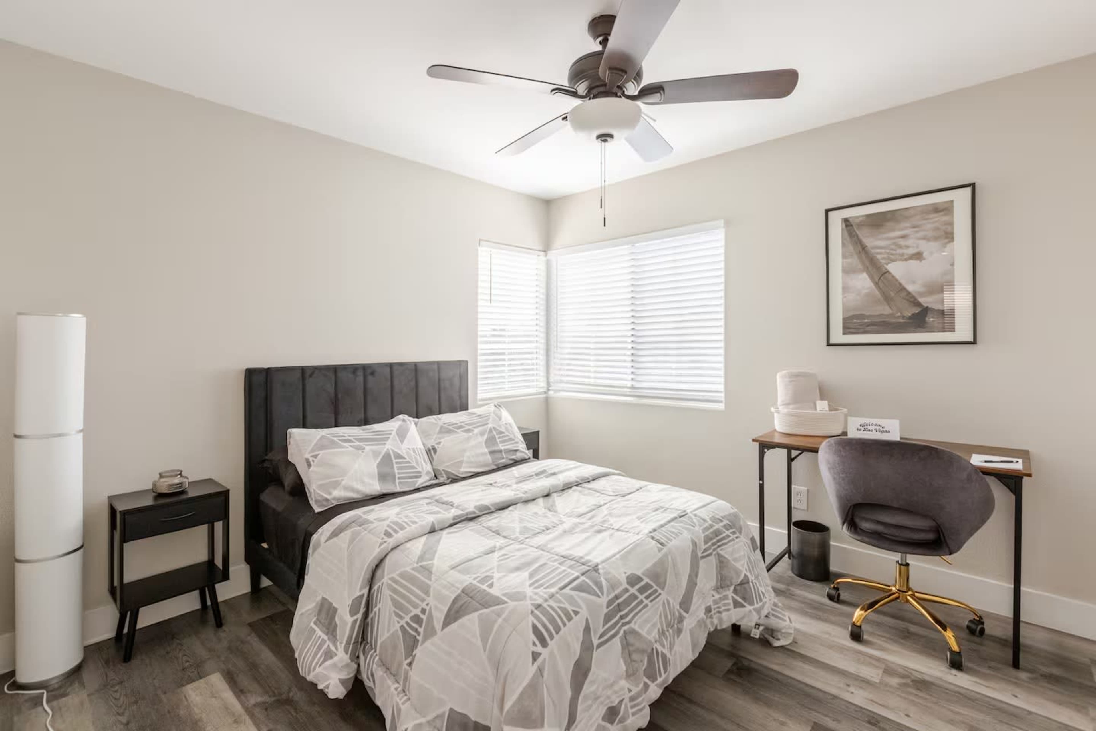 The image shows a small bedroom featuring a bed with a patterned duvet, a desk and chair, and a window with blinds.