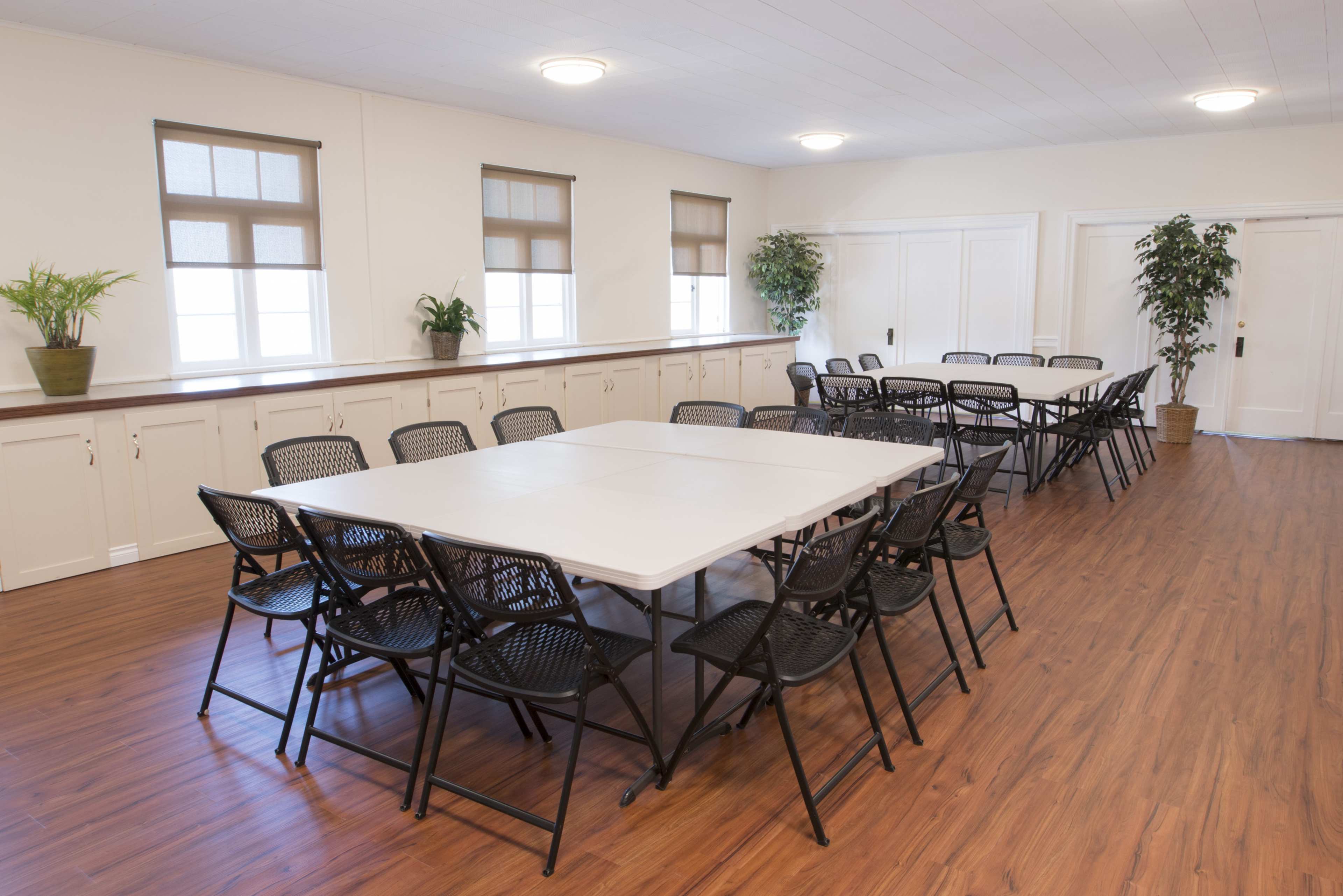 A rectangular arrangement of tables and black chairs occupies a spacious, well-lit room with large windows and potted plants.