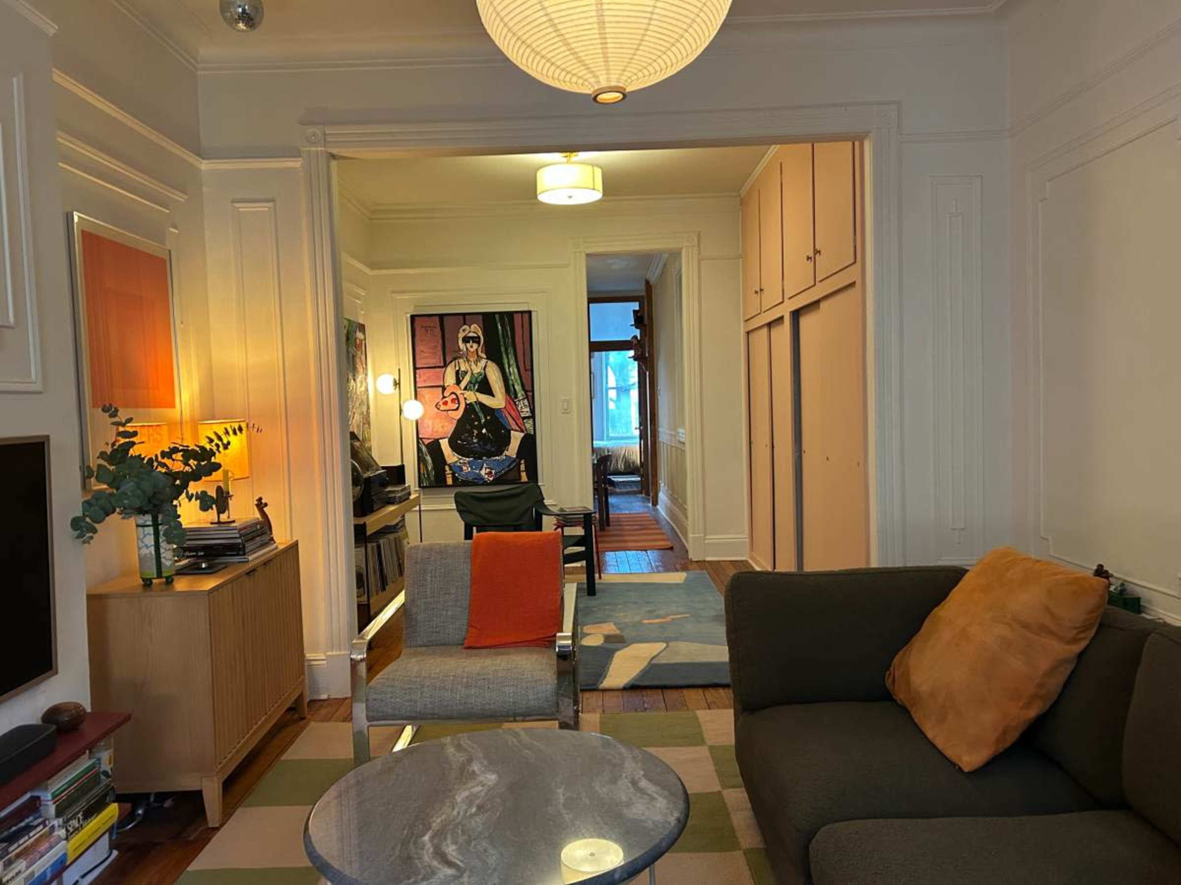 The image shows a living room with a sofa, a coffee table, a lamp, and a hallway leading to another room, featuring artwork on the wall.