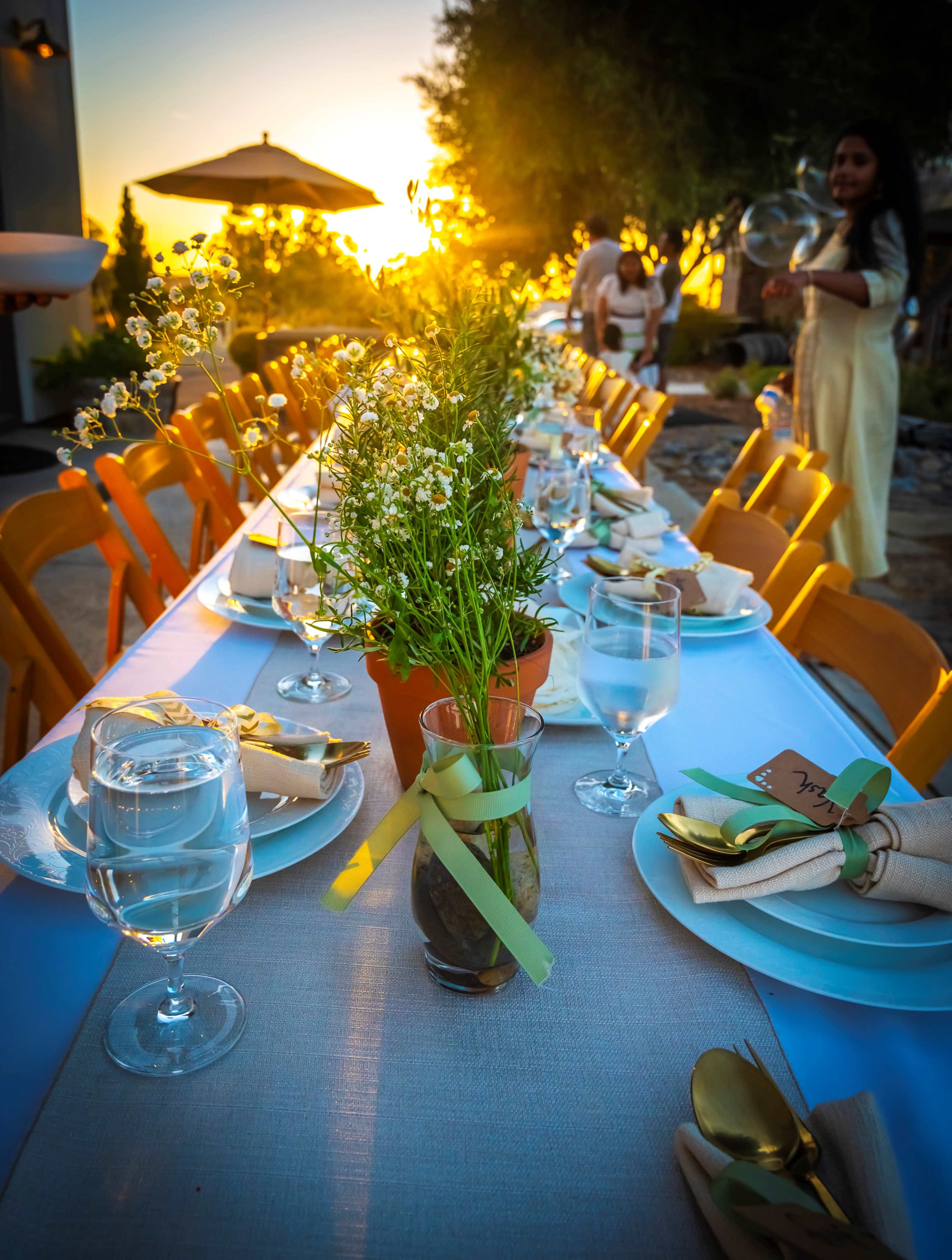 A long table set for an outdoor dinner features flower arrangements and glasses of water, with a sunset in the background.