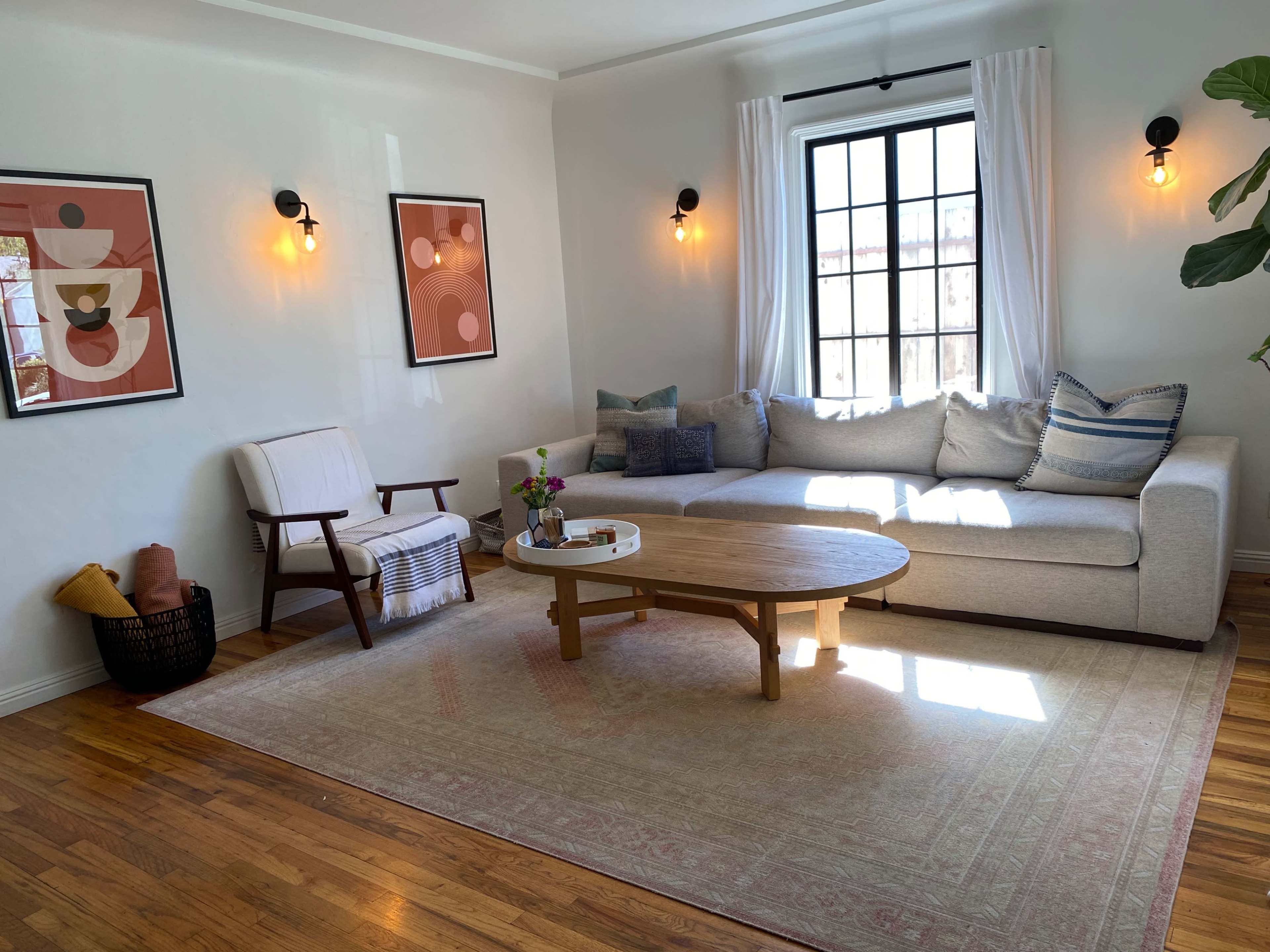 A bright living room featuring a sectional sofa, a wooden coffee table, and decorative wall art, with large windows allowing natural light to fill the space.
