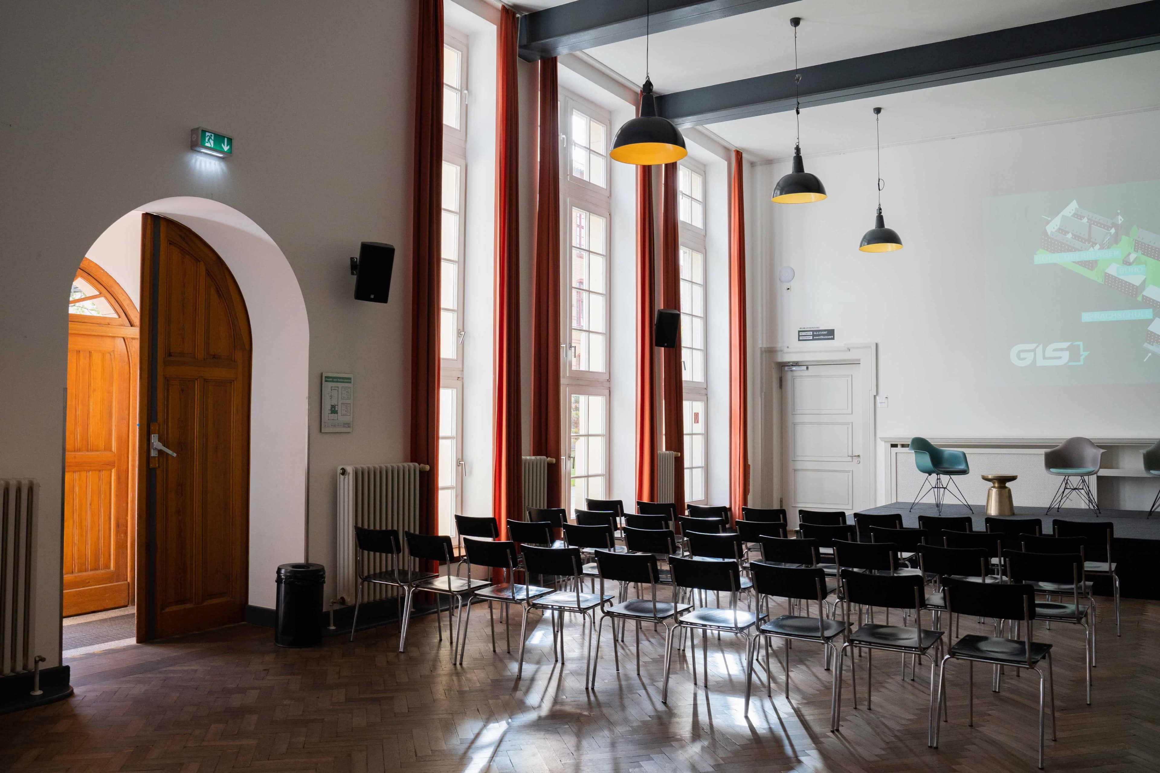 The image shows a seminar room with rows of black chairs facing a blank screen, large windows on the walls, and a wooden door leading to another area.