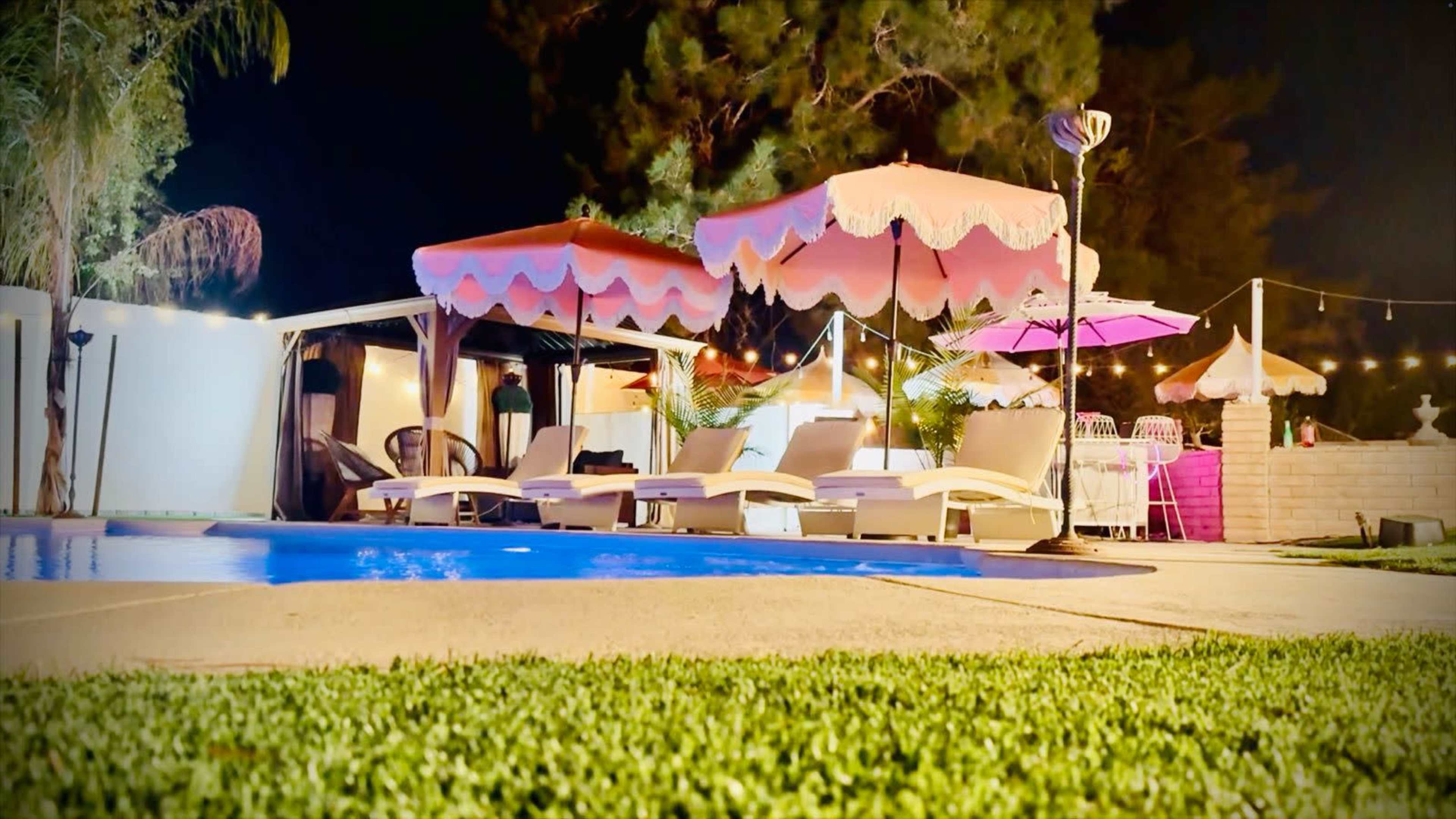 The scene features a nighttime pool area with pink umbrellas over lounge chairs, surrounded by lush green grass and softly lit by string lights.
