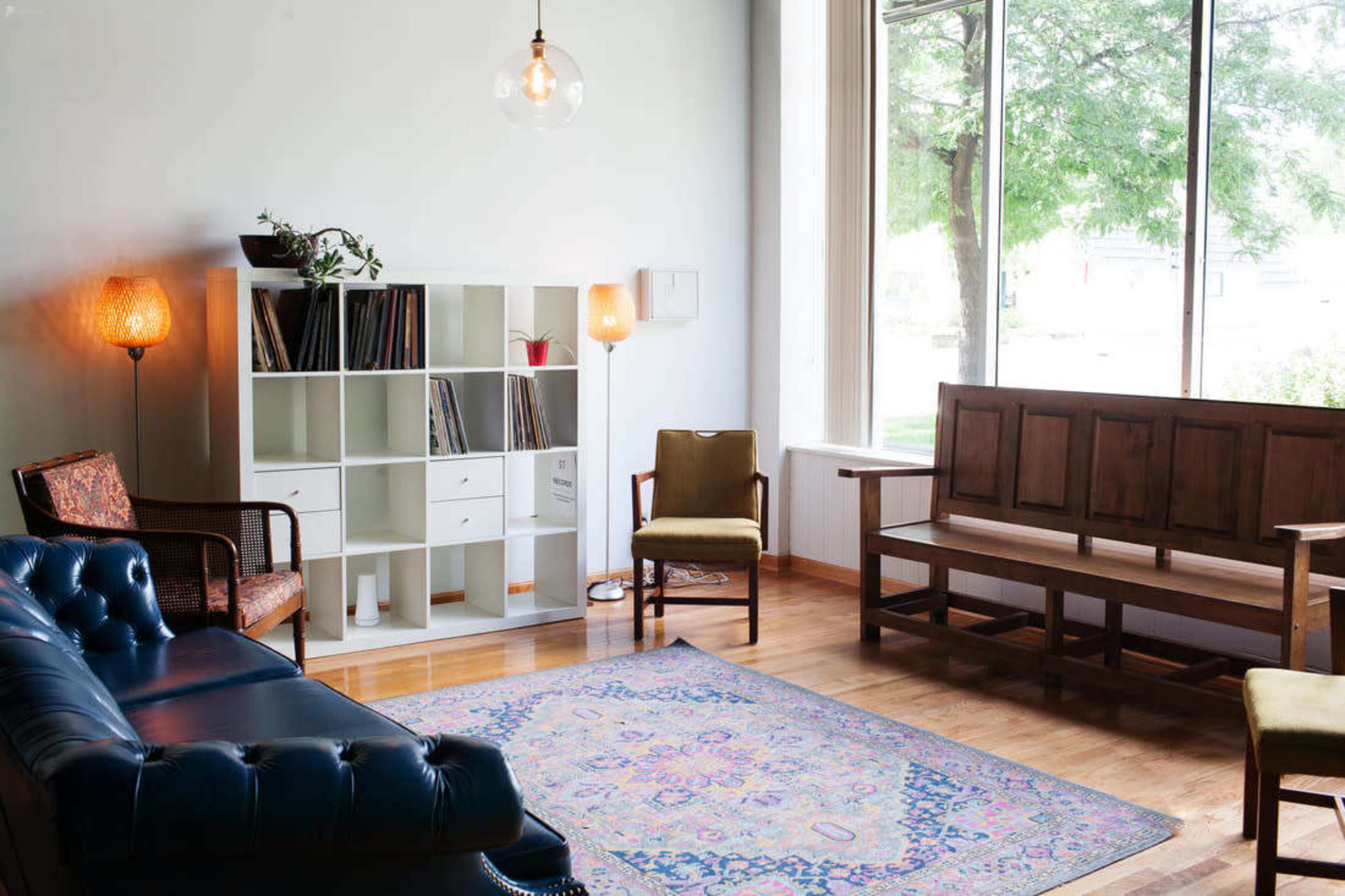 A well-lit living room features a blue leather sofa, a wooden bench, a shelving unit filled with records, and a large window revealing greenery outside.