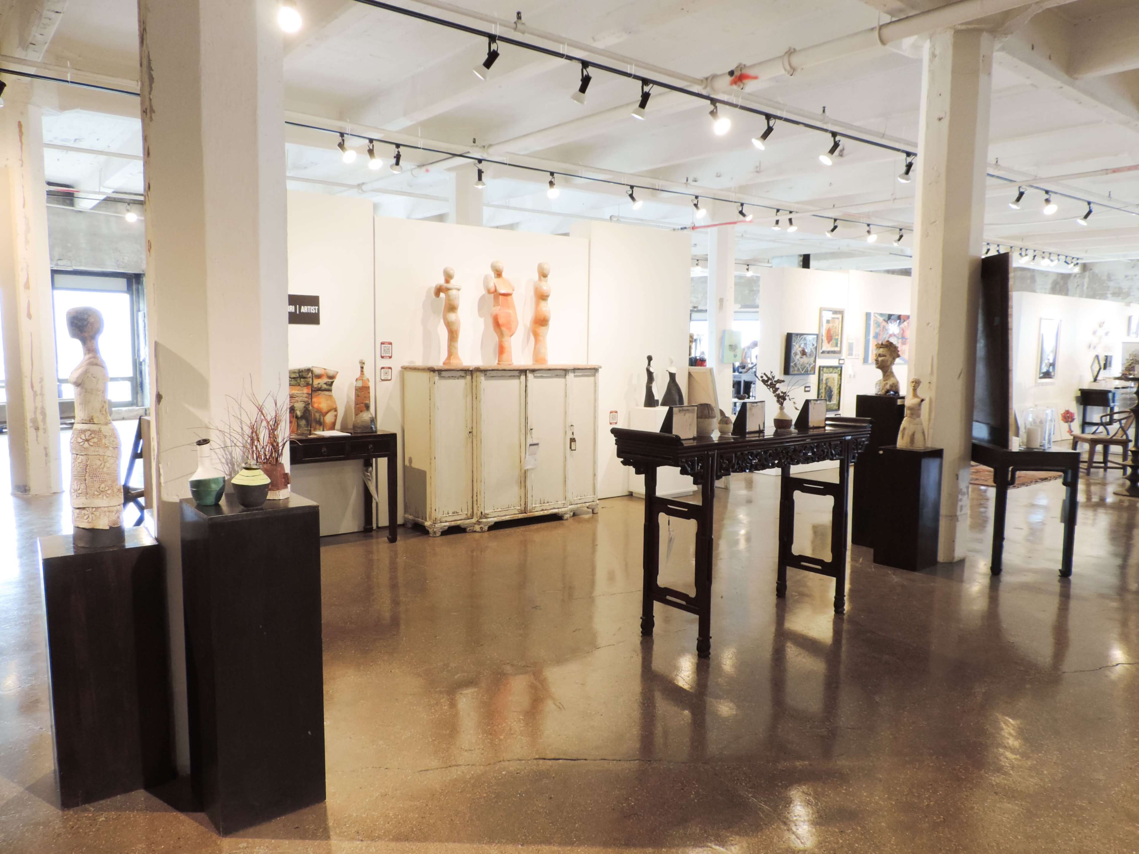 The image shows an art gallery featuring various sculptures and furniture displayed in a spacious, well-lit room with polished concrete floors.
