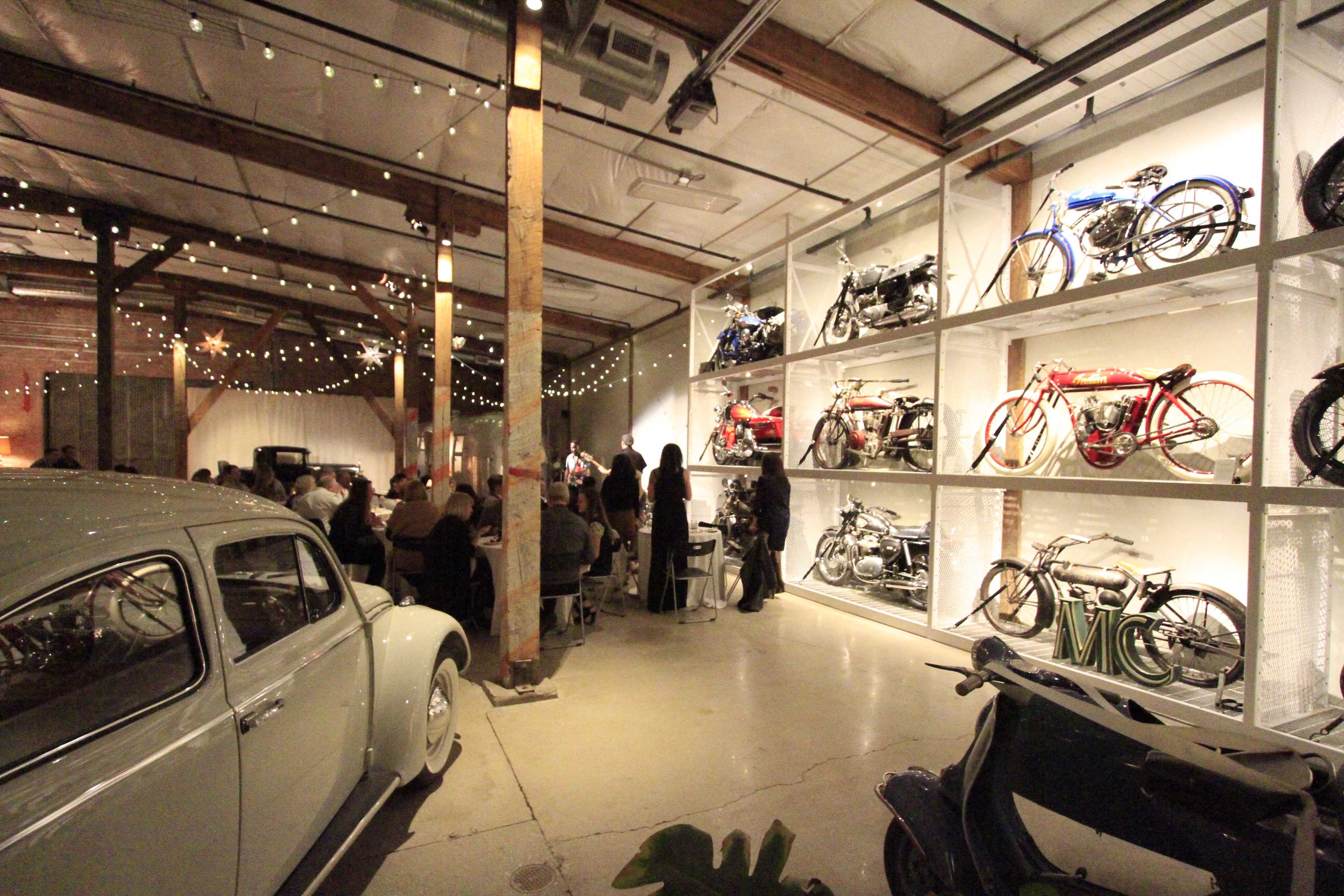 A large indoor space with decorative string lights, featuring vintage bicycles displayed on wall shelves alongside a classic car and tables where people are seated.