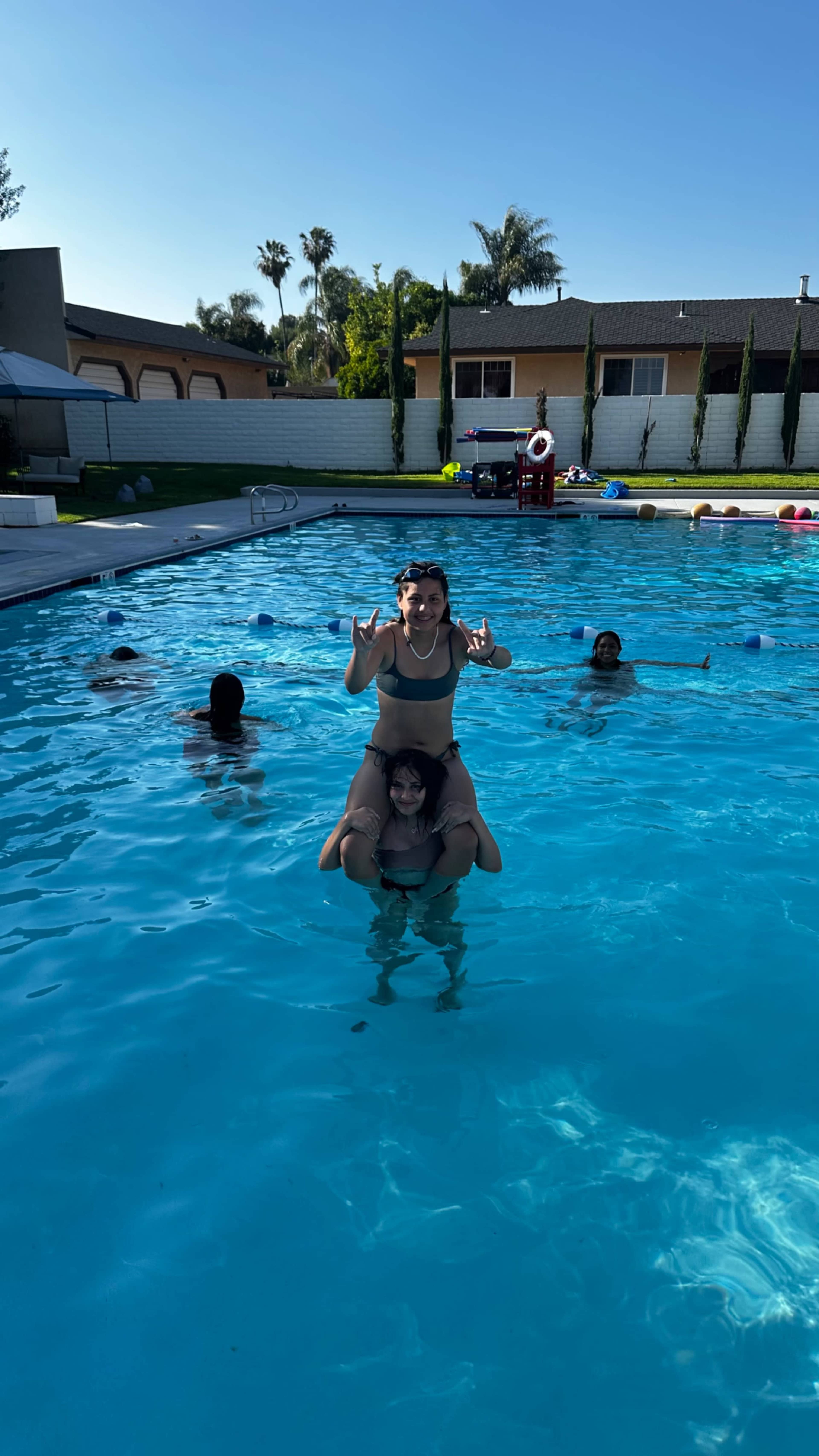 A group of children is playing in a swimming pool, with two kids performing a playful act where one is standing on the shoulders of another.