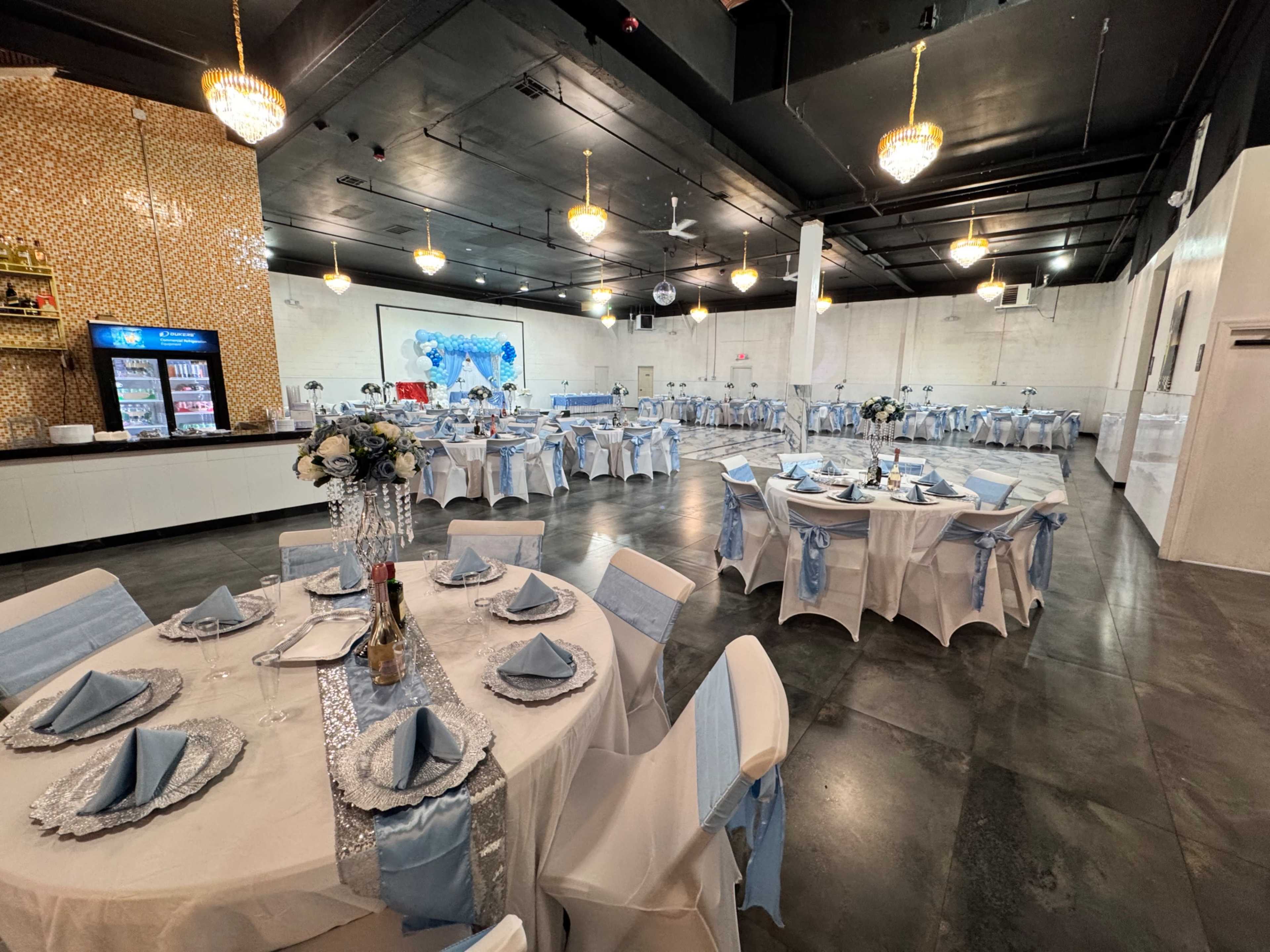 The image shows a banquet hall with white tables covered in light blue tablecloths and set for an event, featuring a bar area and decorative elements on the walls.
