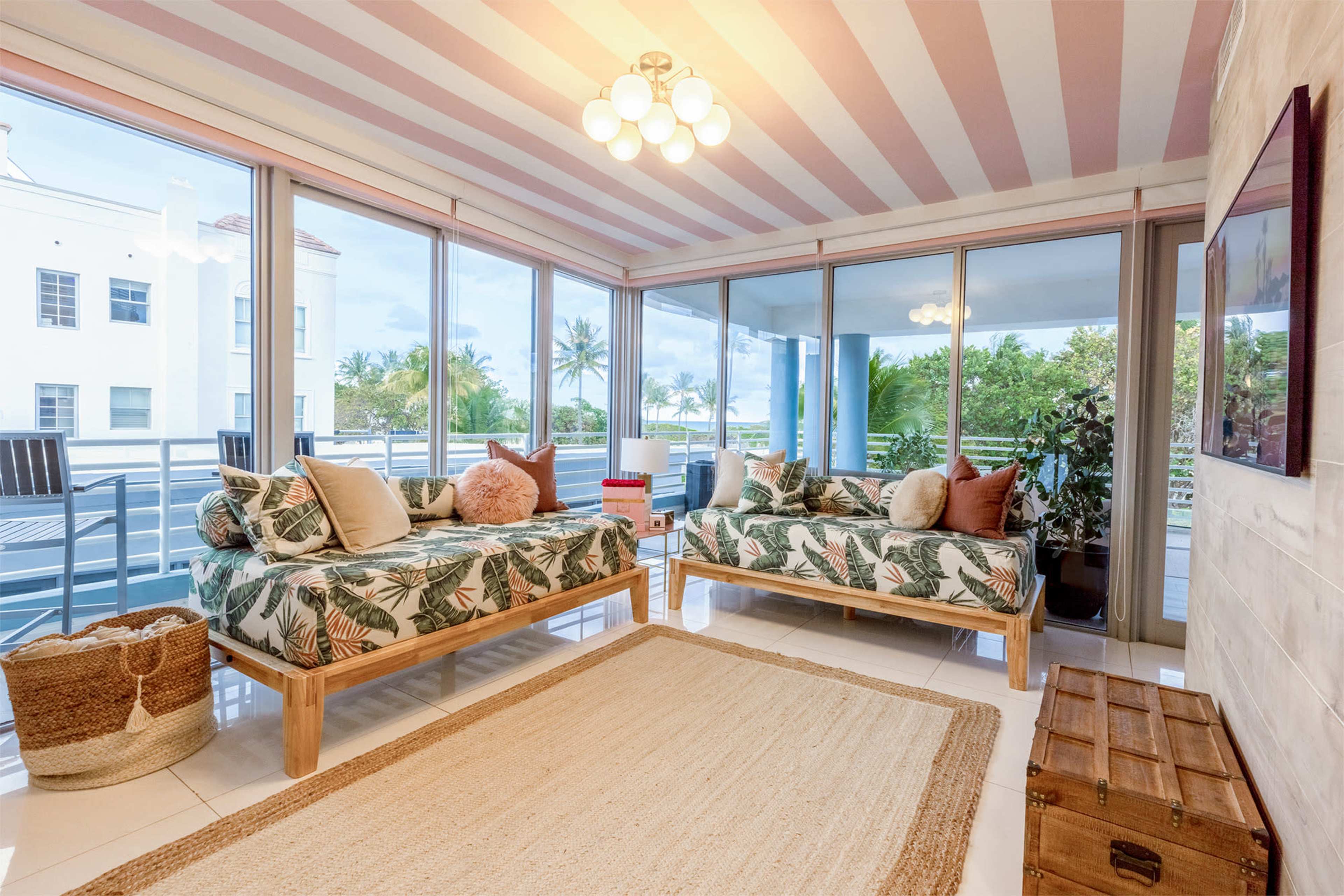 A bright living space features two beds with tropical-patterned bedding, large windows overlooking palm trees, and a light-colored area rug on a glossy floor.