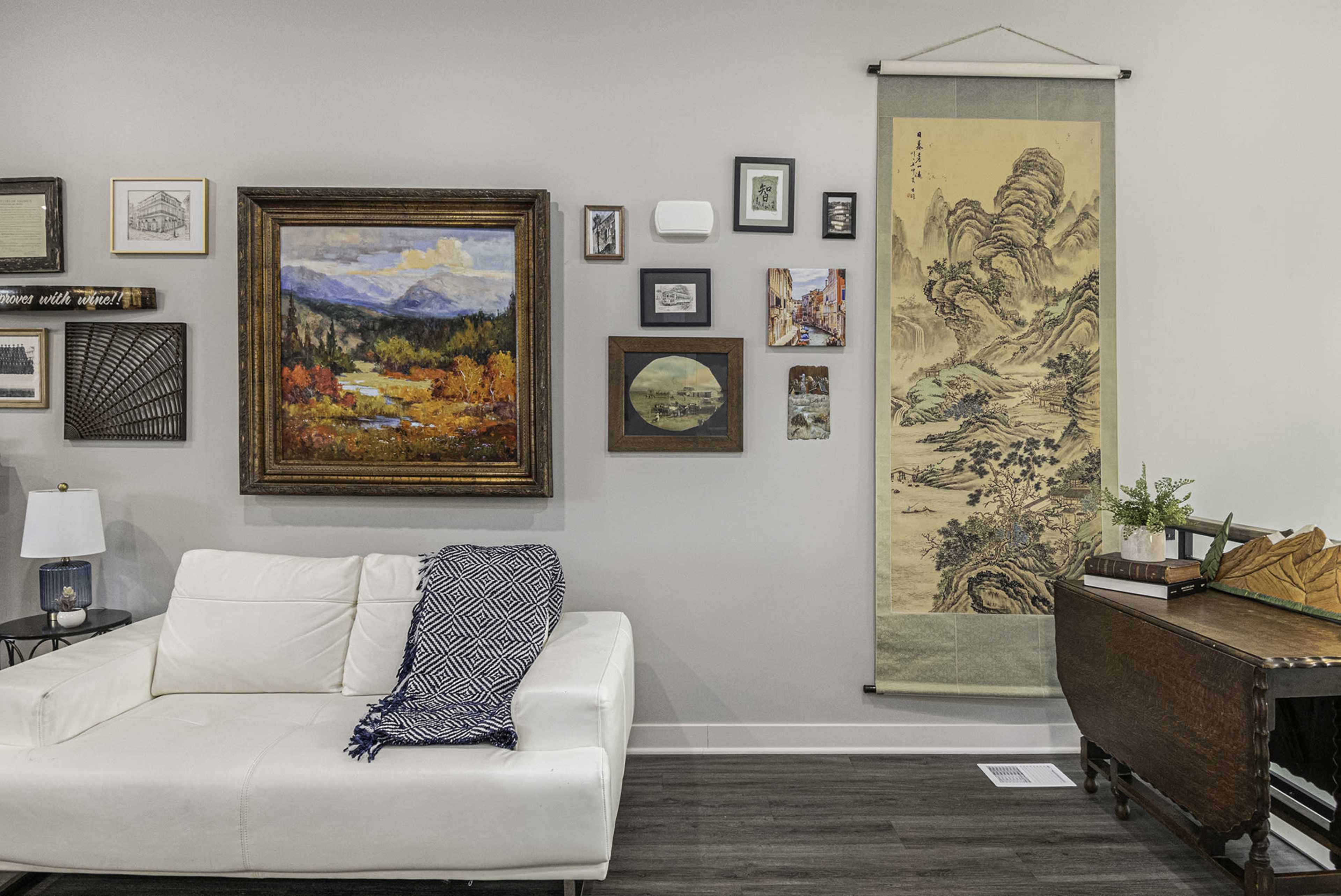 A living room wall adorned with various framed artworks, including a landscape painting, alongside a hanging scroll, with a white couch and a small table in the foreground.