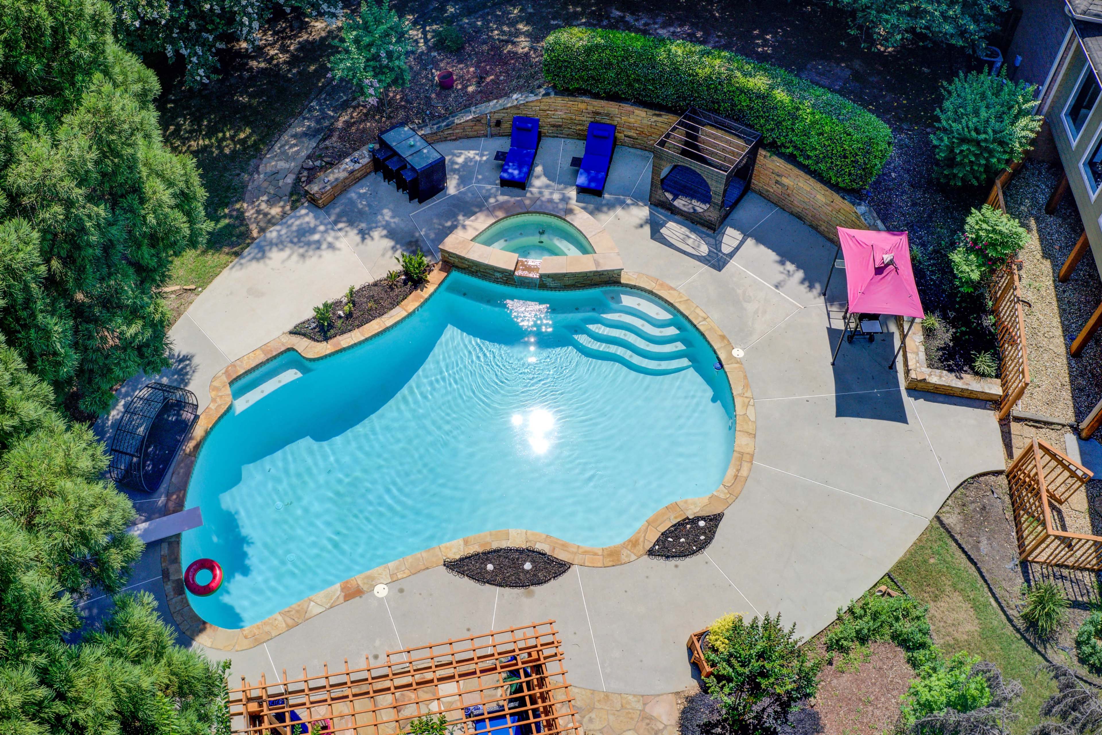 The image shows an aerial view of a curved swimming pool surrounded by patio furniture and landscaped greenery.