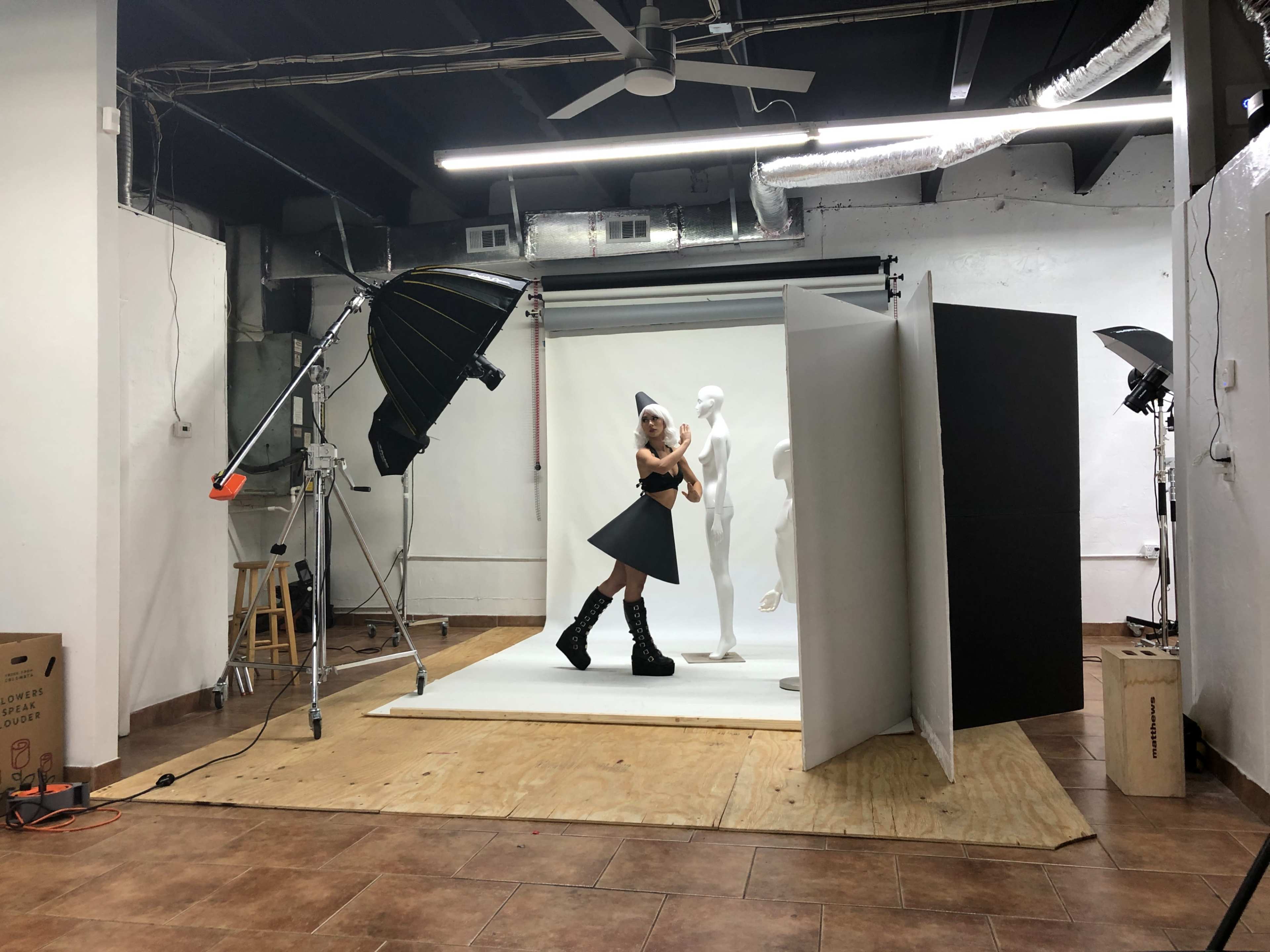 A model poses in front of a white backdrop in a photography studio, surrounded by lighting equipment and a mannequin.