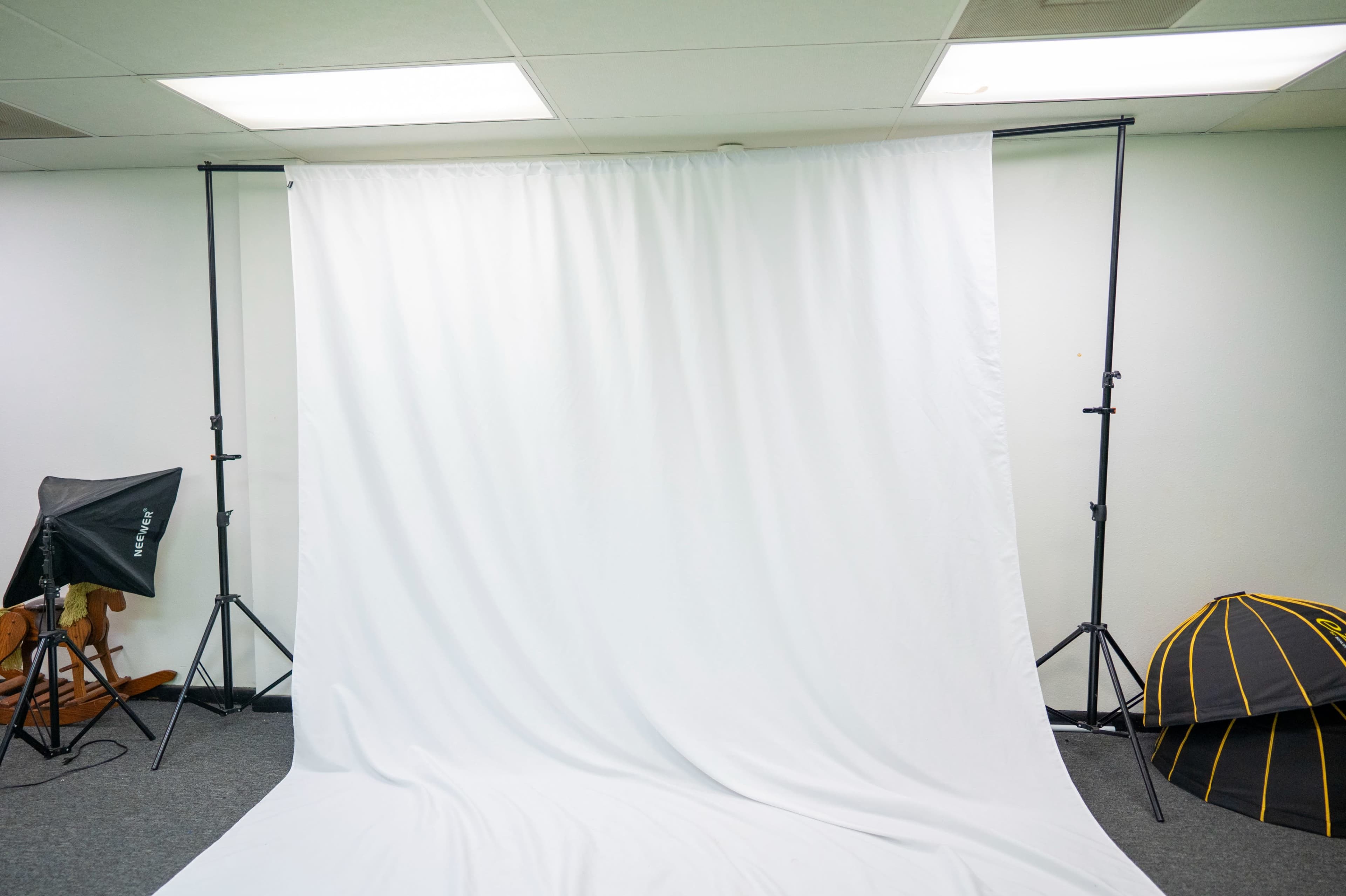 The image shows a plain white backdrop setup on a stand in a room, with studio lighting equipment and a folded black and yellow object in the corner.