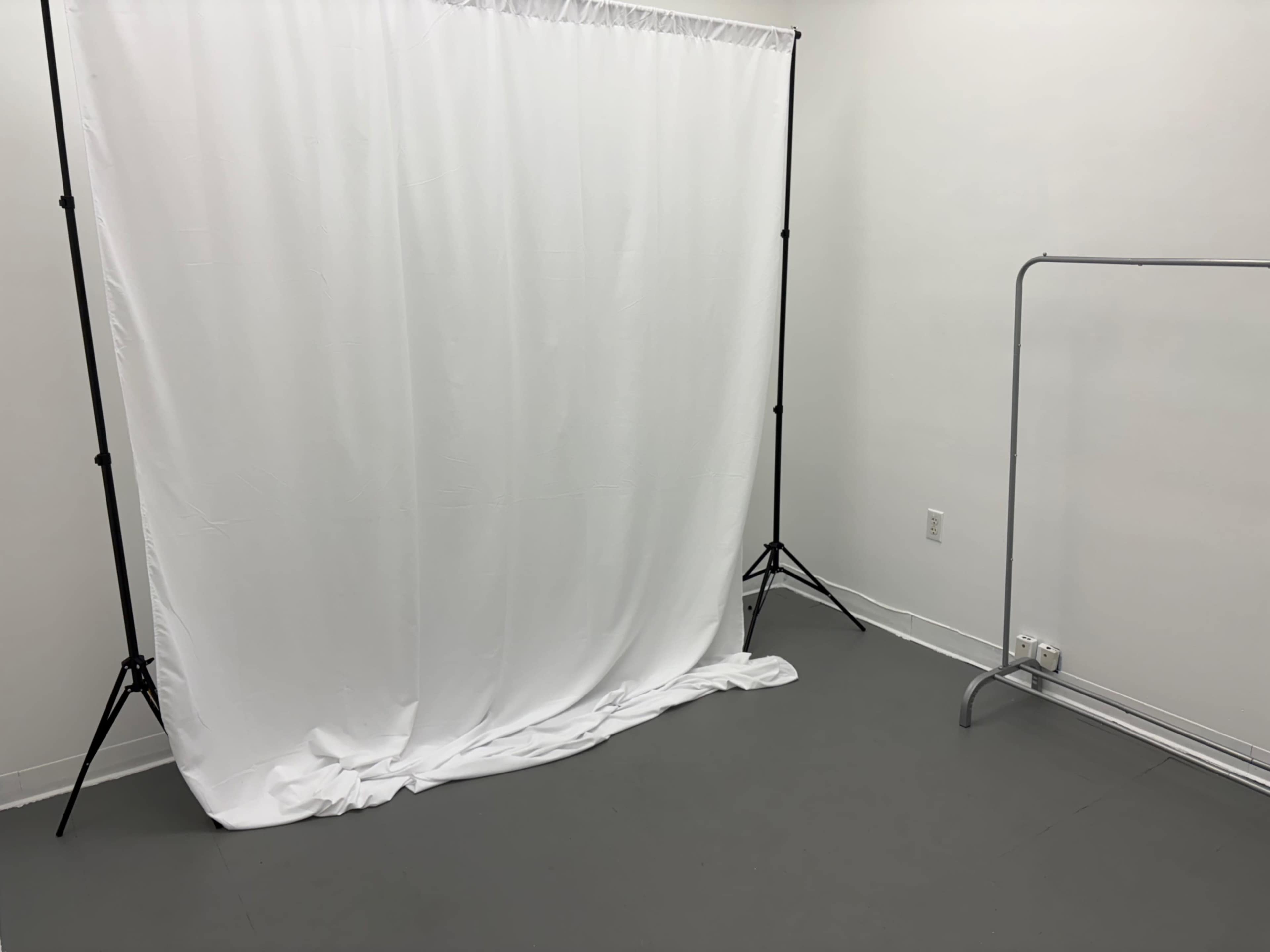 The image shows an empty room with a white backdrop draped on a stand and a clothing rack in the corner.