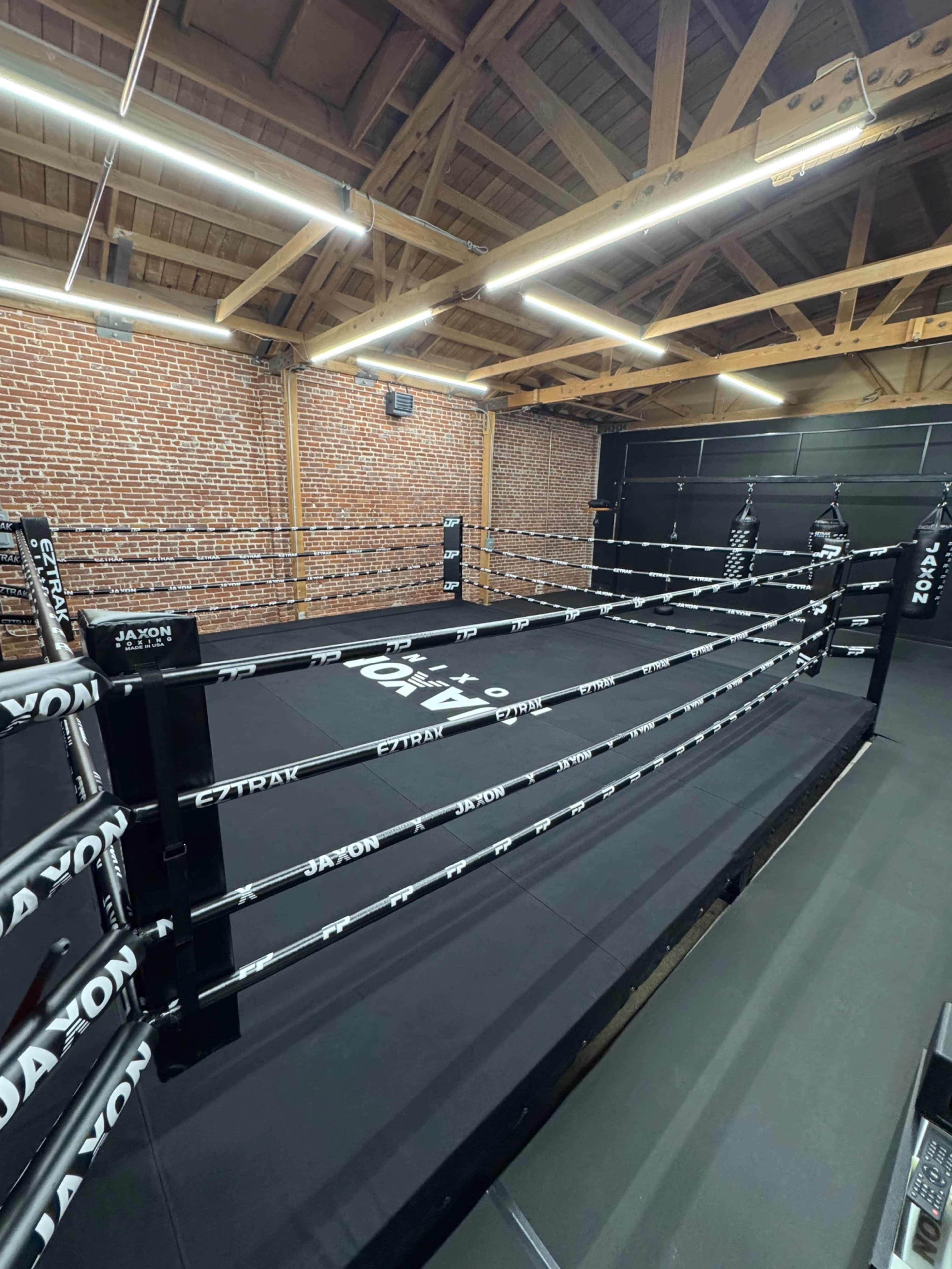 The image shows a boxing gym featuring a boxing ring with black padding and white ropes, surrounded by exposed brick walls and wooden beams.