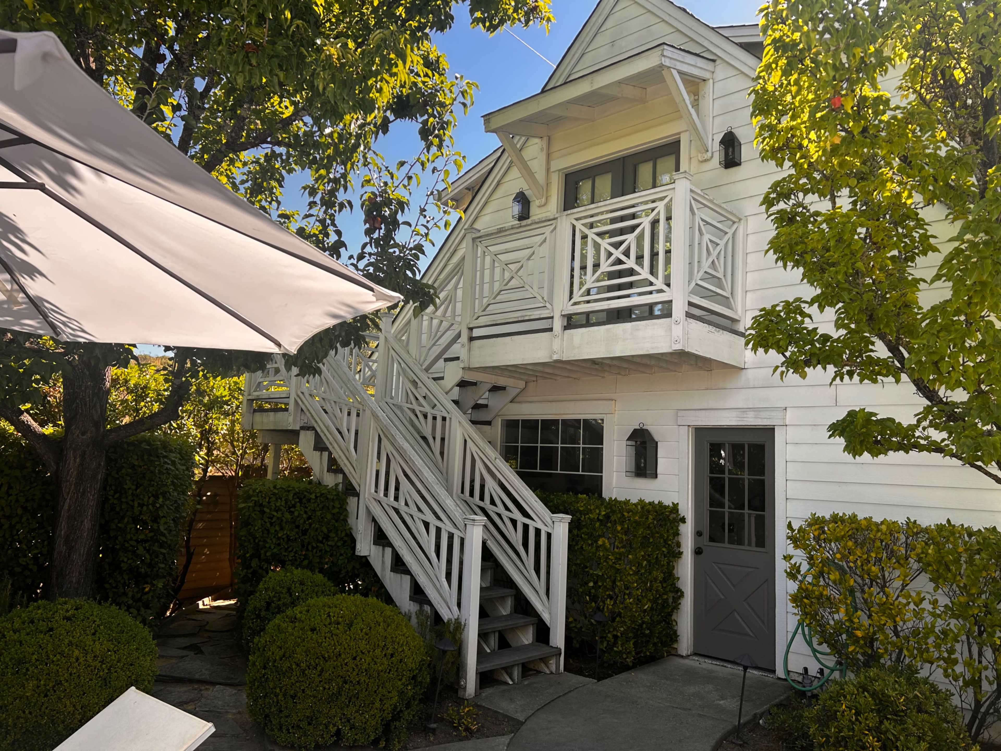 A two-story white house with a front staircase and balcony is surrounded by neatly trimmed bushes and trees.