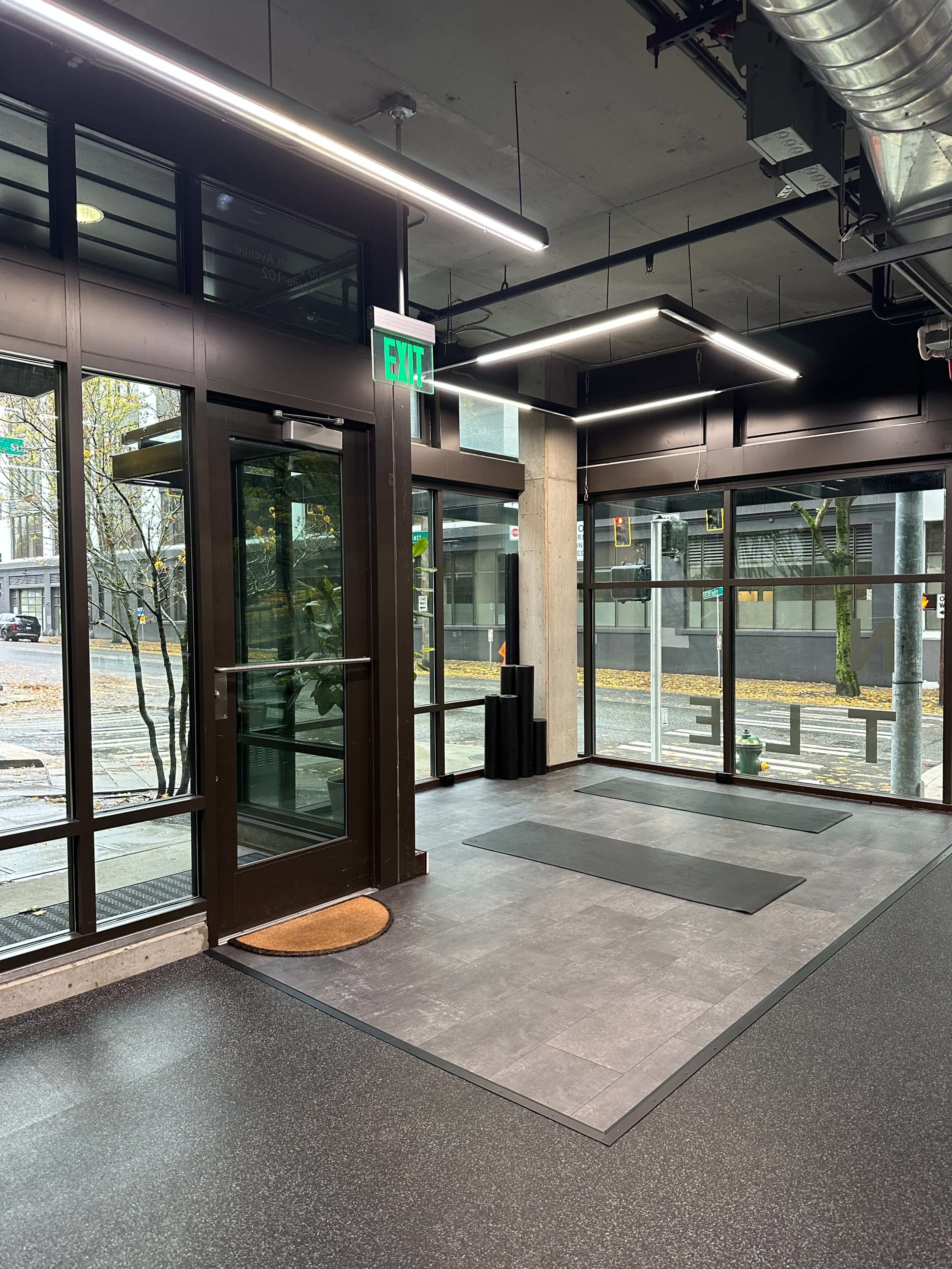 The image shows a modern interior space with large glass windows, an exit door, and mats on the floor, indicating a fitness or yoga area.