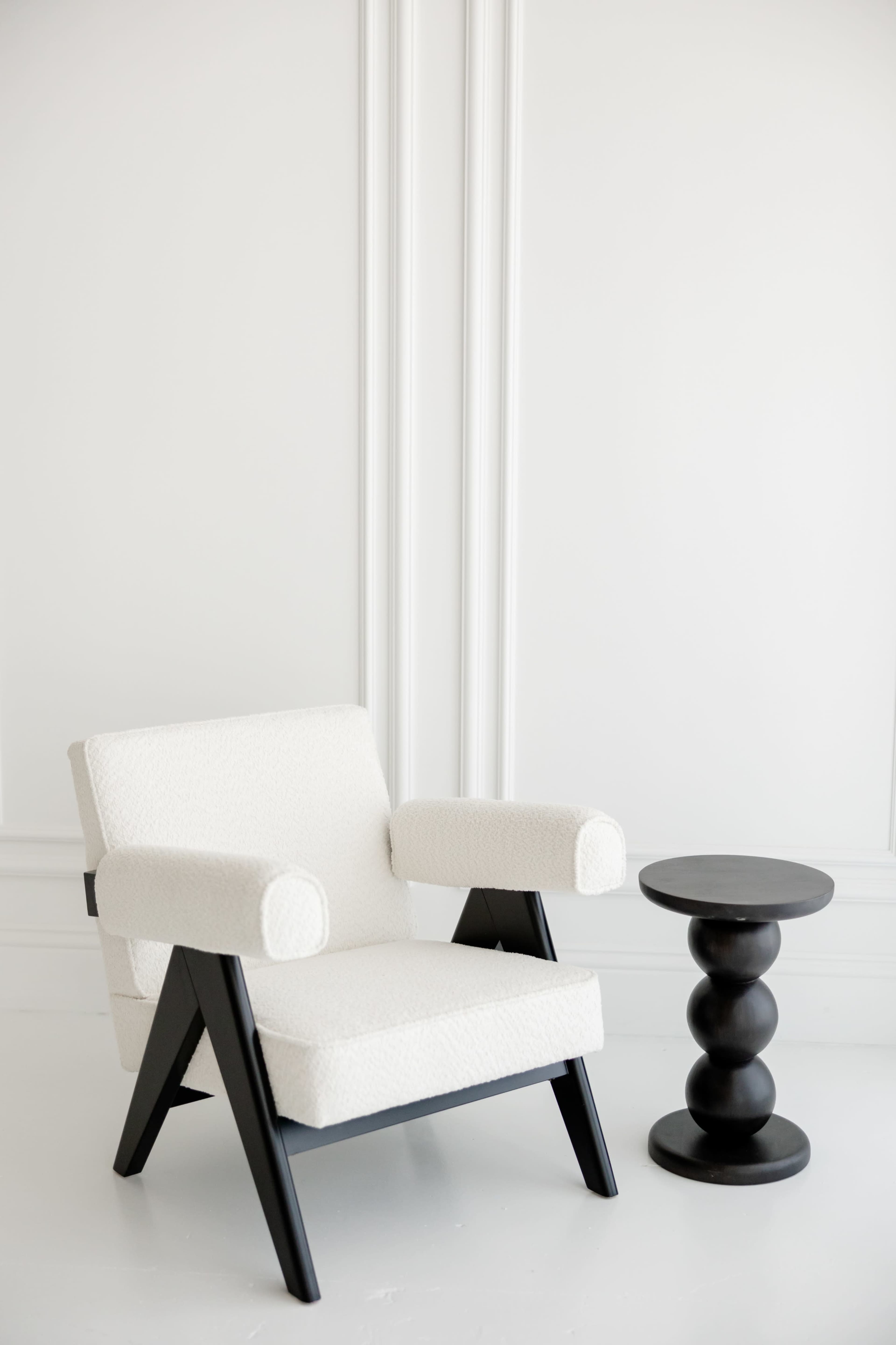 A white upholstered armchair with black wooden legs sits next to a black, round side table in a minimalist room.