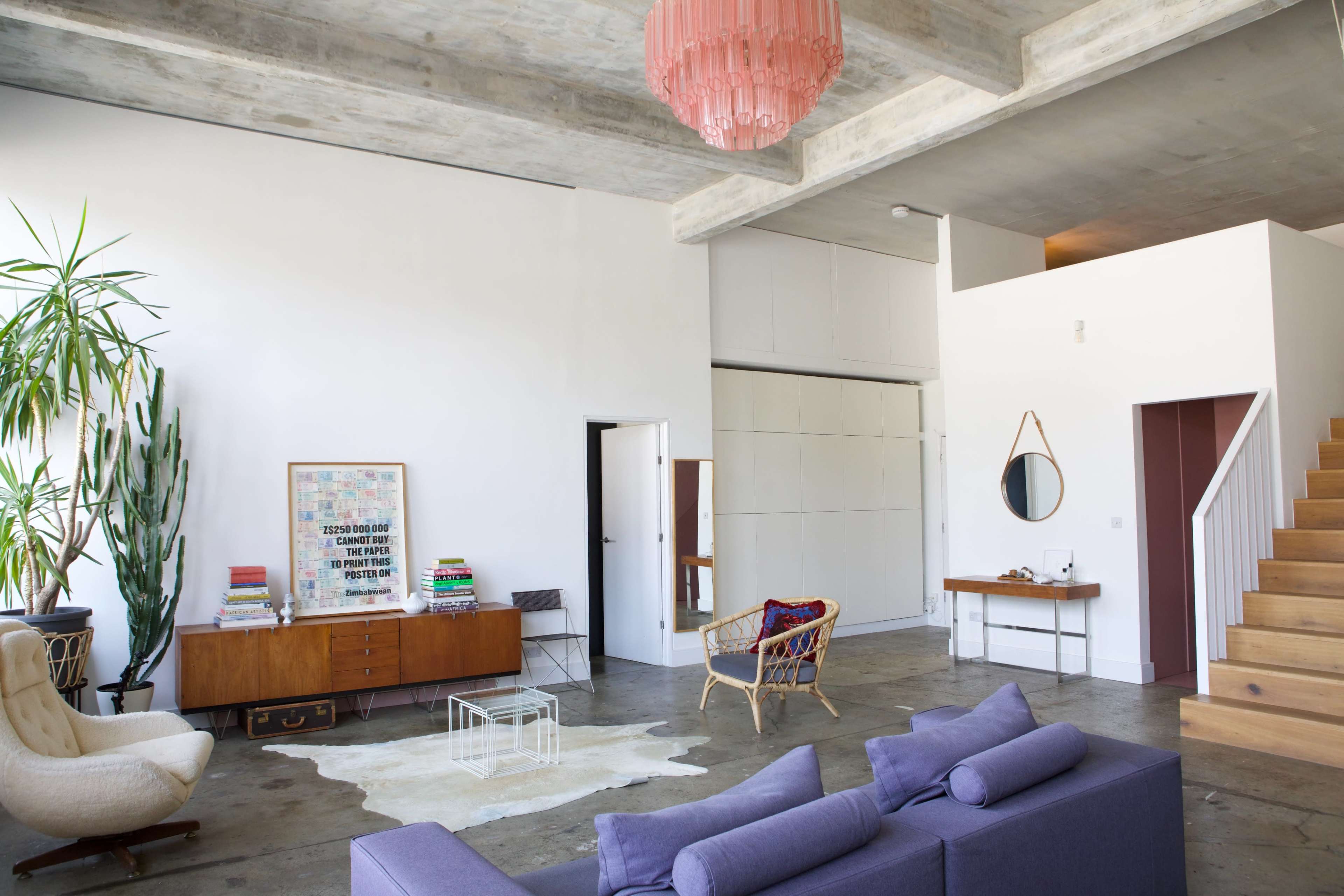The image features a modern living room with a large gray sofa, a decorative rug on a polished concrete floor, a tall indoor plant, and a staircase leading to an upper level.