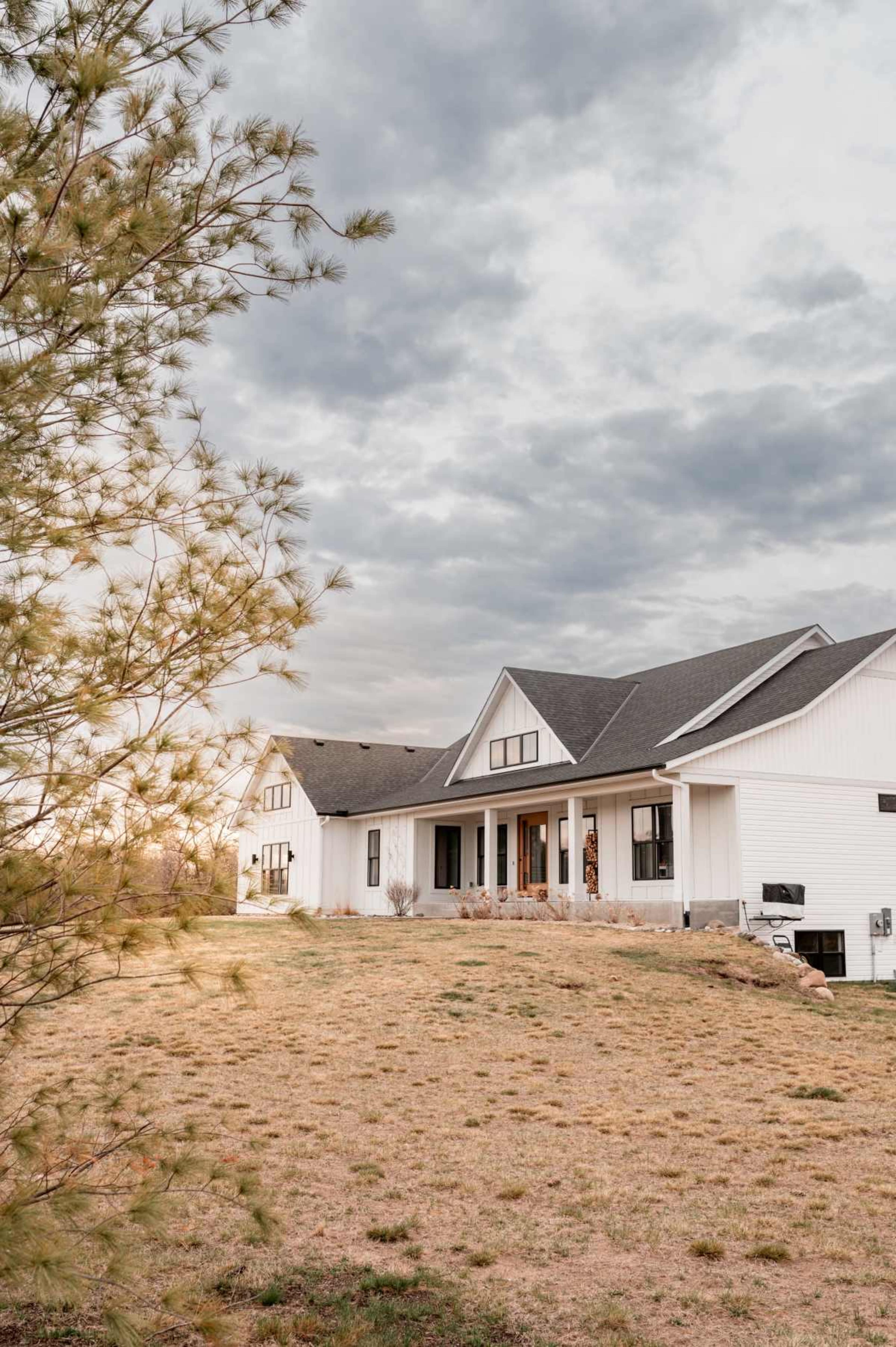 A modern white house with a gray roof sits on a grassy hill under a cloudy sky.