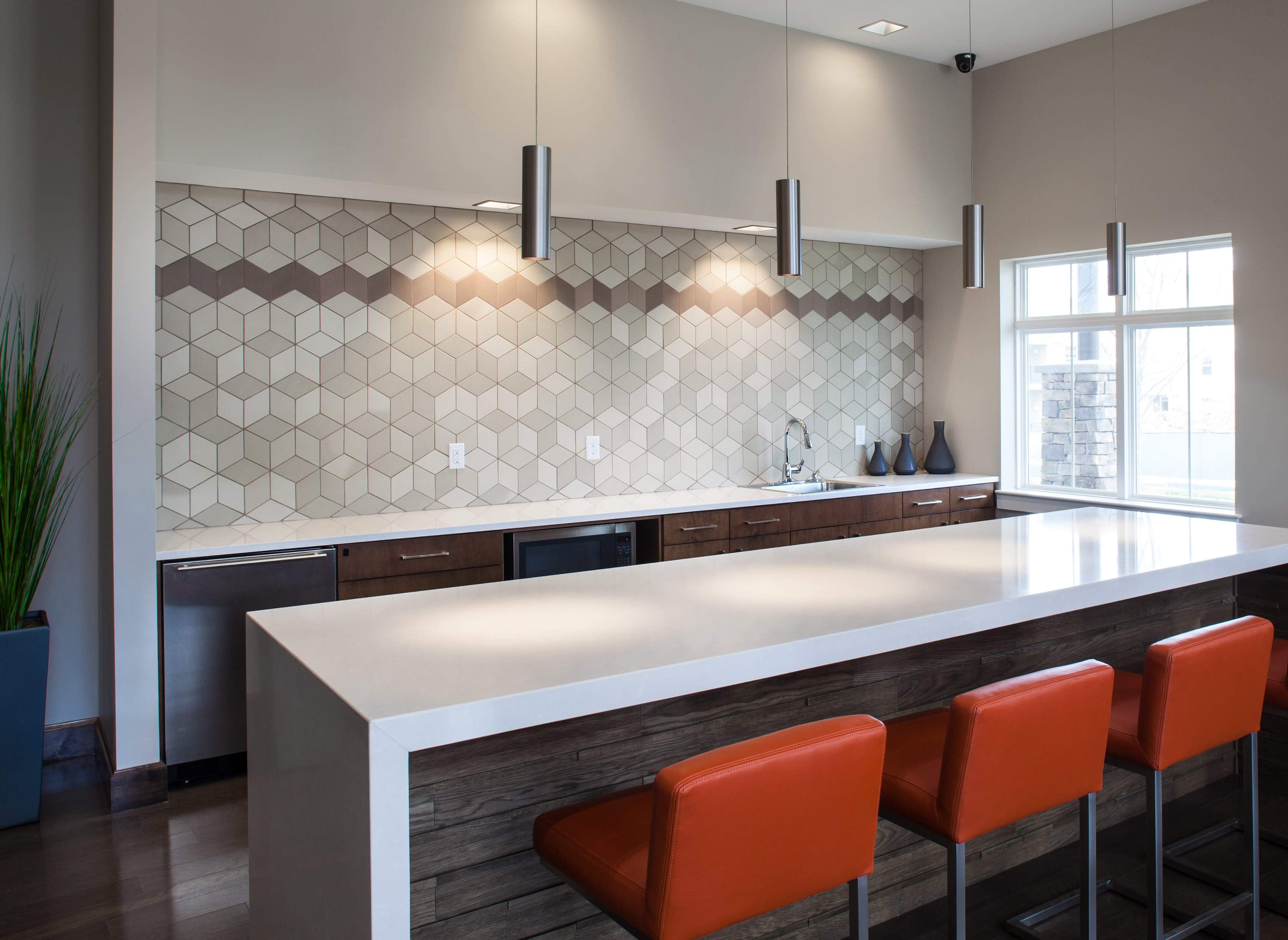 A modern kitchen features a hexagonal tiled backsplash, a long white countertop, and orange stools at the island.