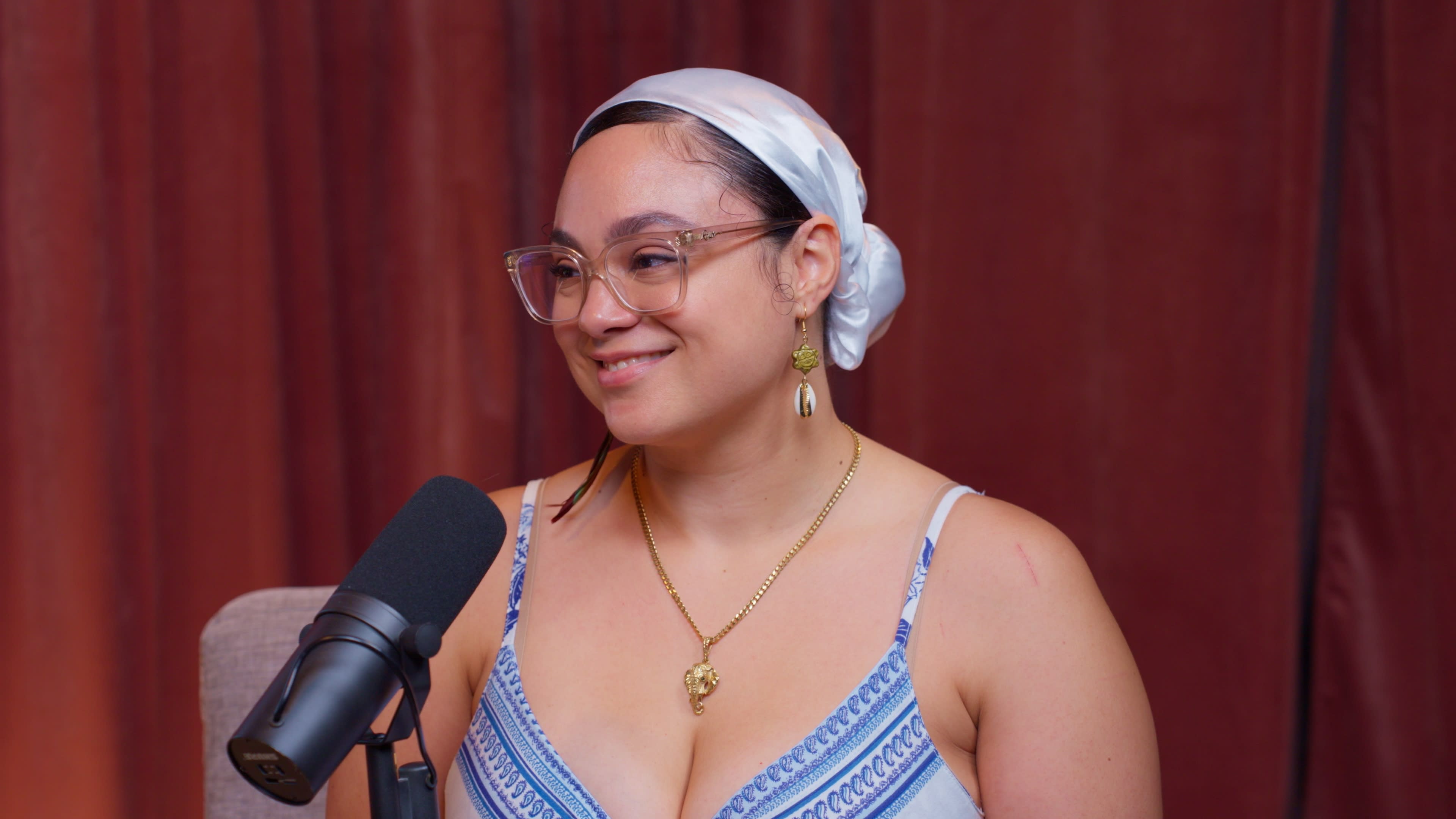 A woman with glasses and a headscarf smiles while seated next to a microphone.