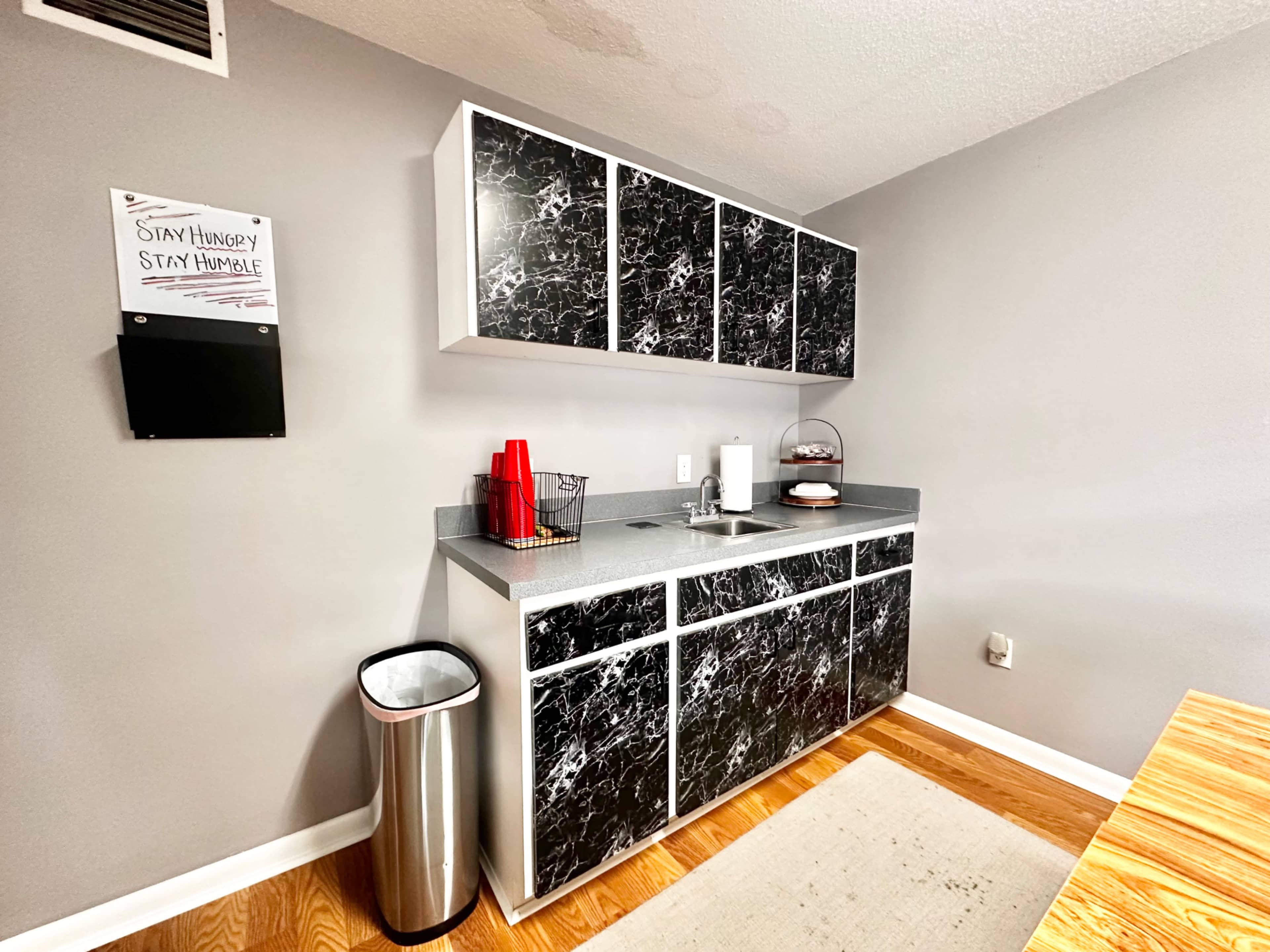 The image shows a compact kitchen area with black marble-patterned cabinets, a sink, and a countertop, alongside a trash can and a motivational quote on the wall.