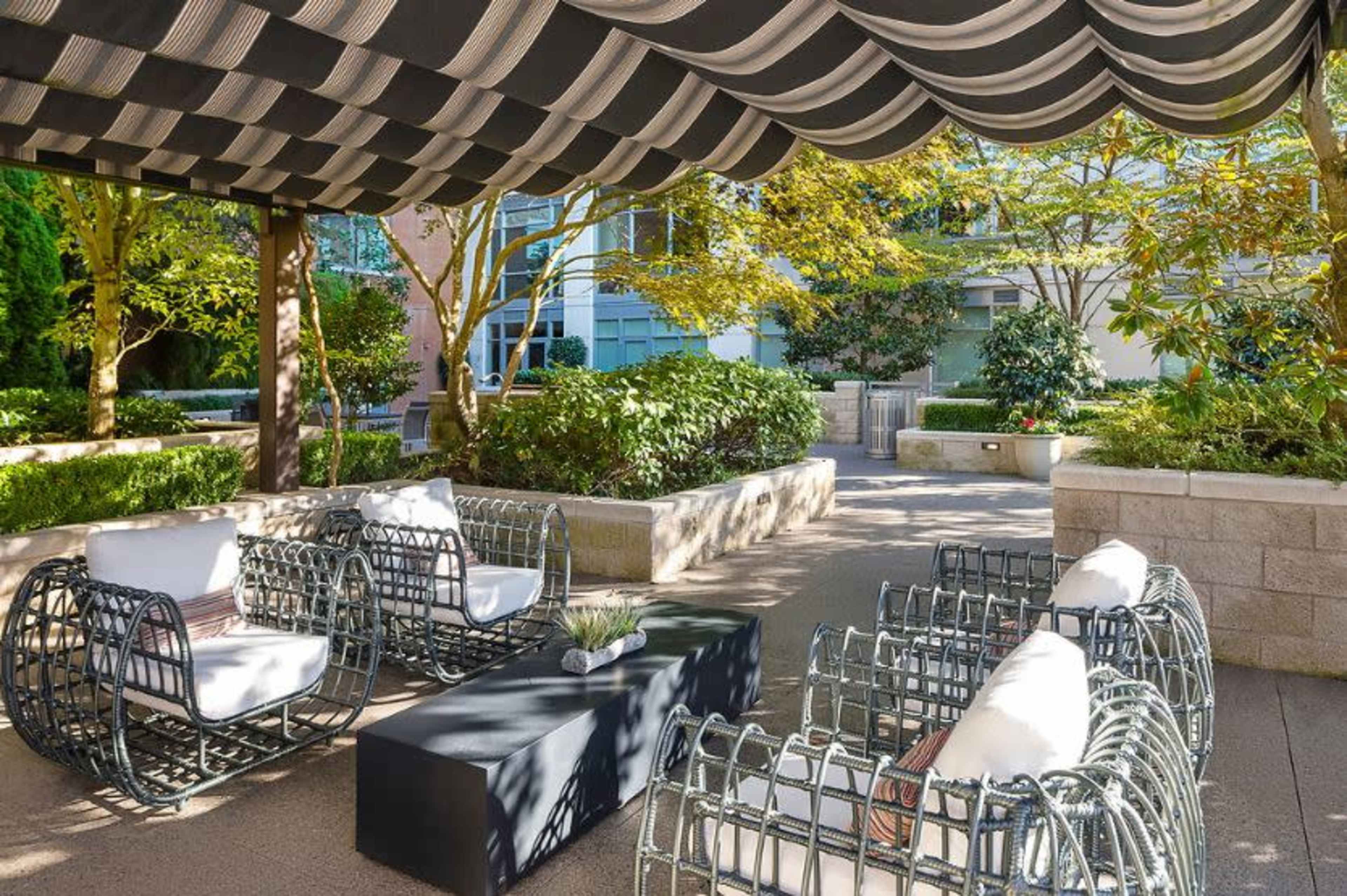 A seating area featuring wire-framed chairs and a table, sheltered under a striped awning amid landscaped greenery.