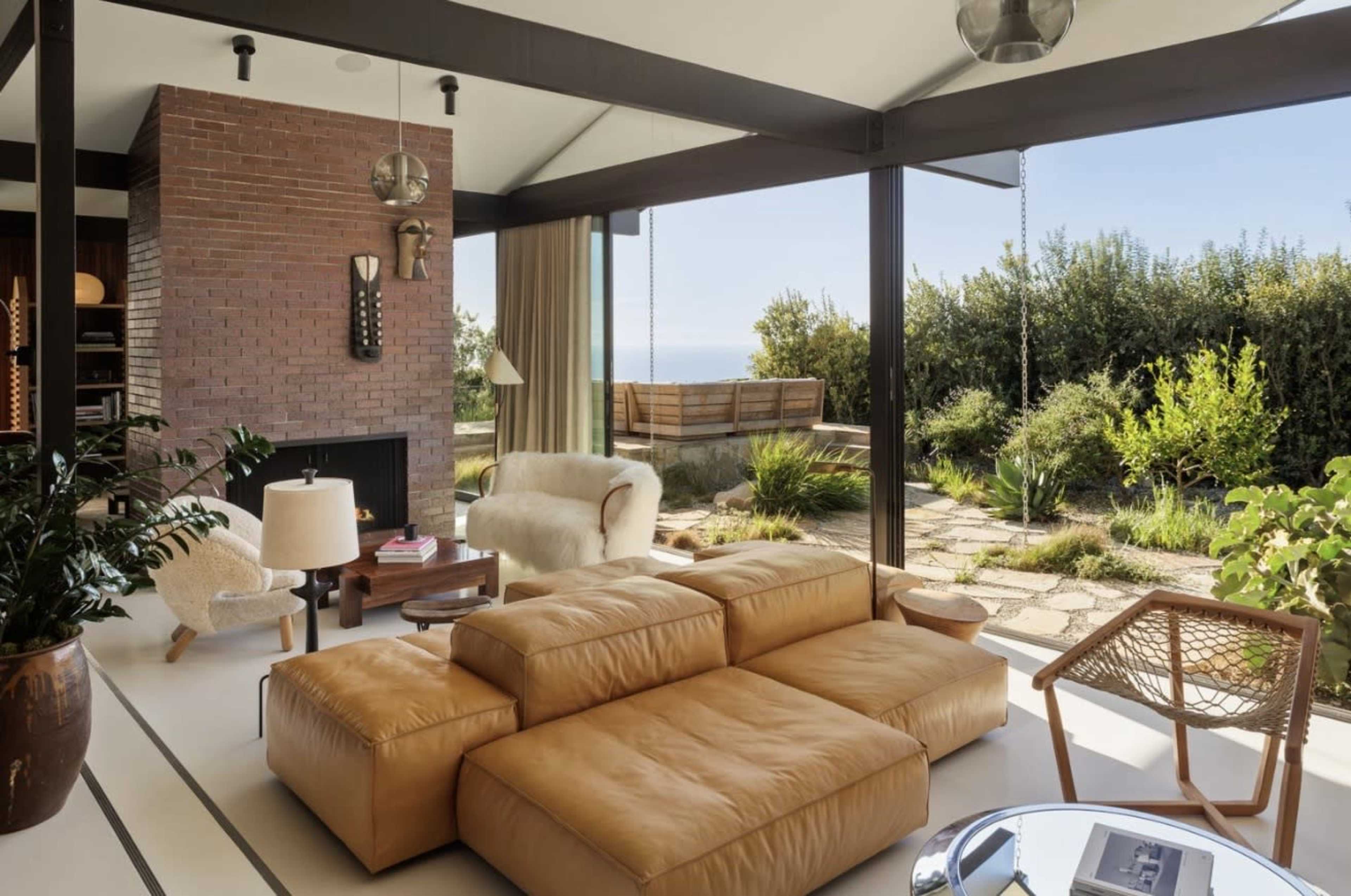 A modern living room features a large leather sofa, a cozy armchair, a coffee table, and expansive glass doors that open to a garden overlooking the ocean.