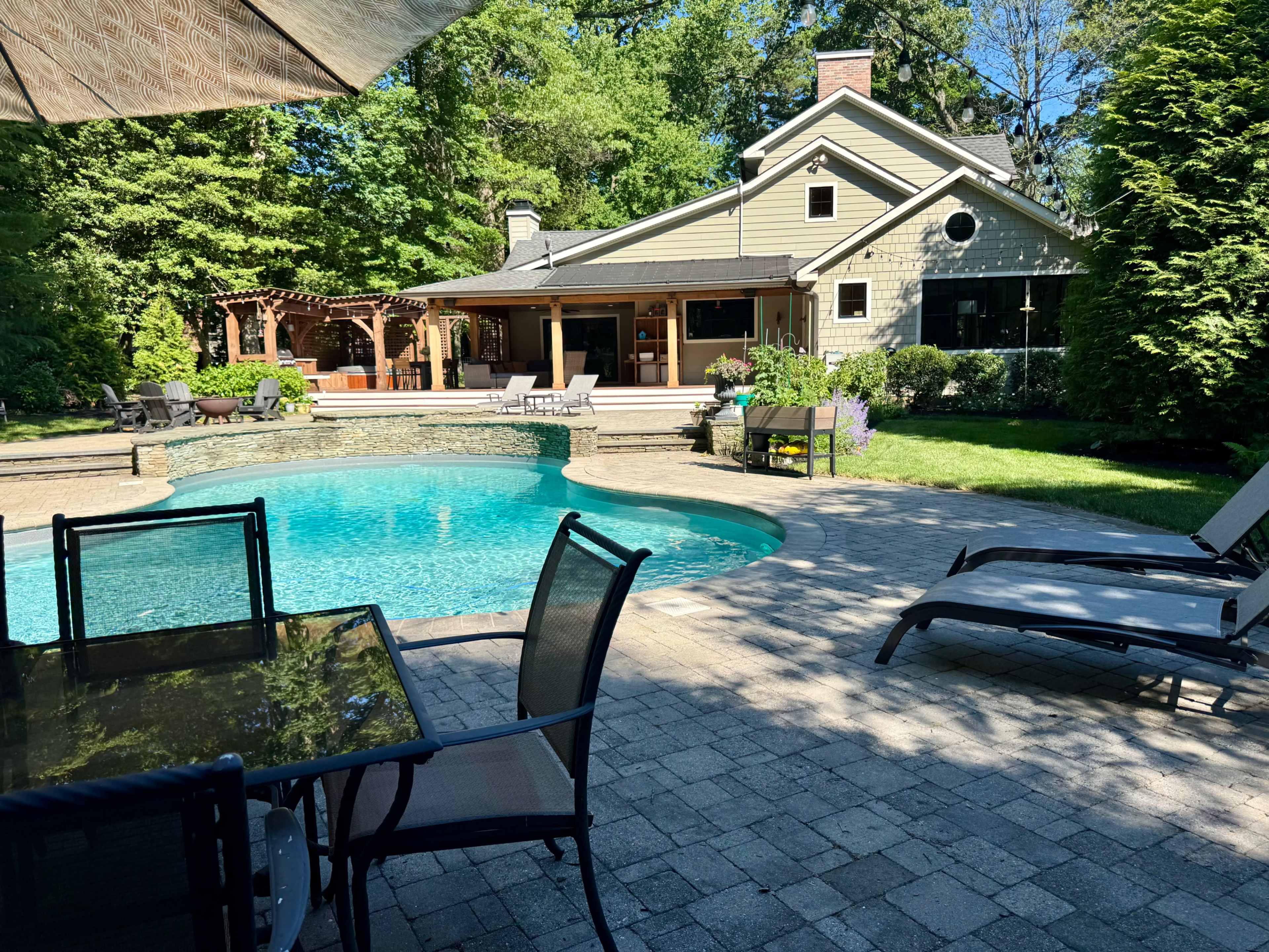 The image shows a landscaped backyard featuring a blue swimming pool, outdoor seating, and a view of a house with a patio area.