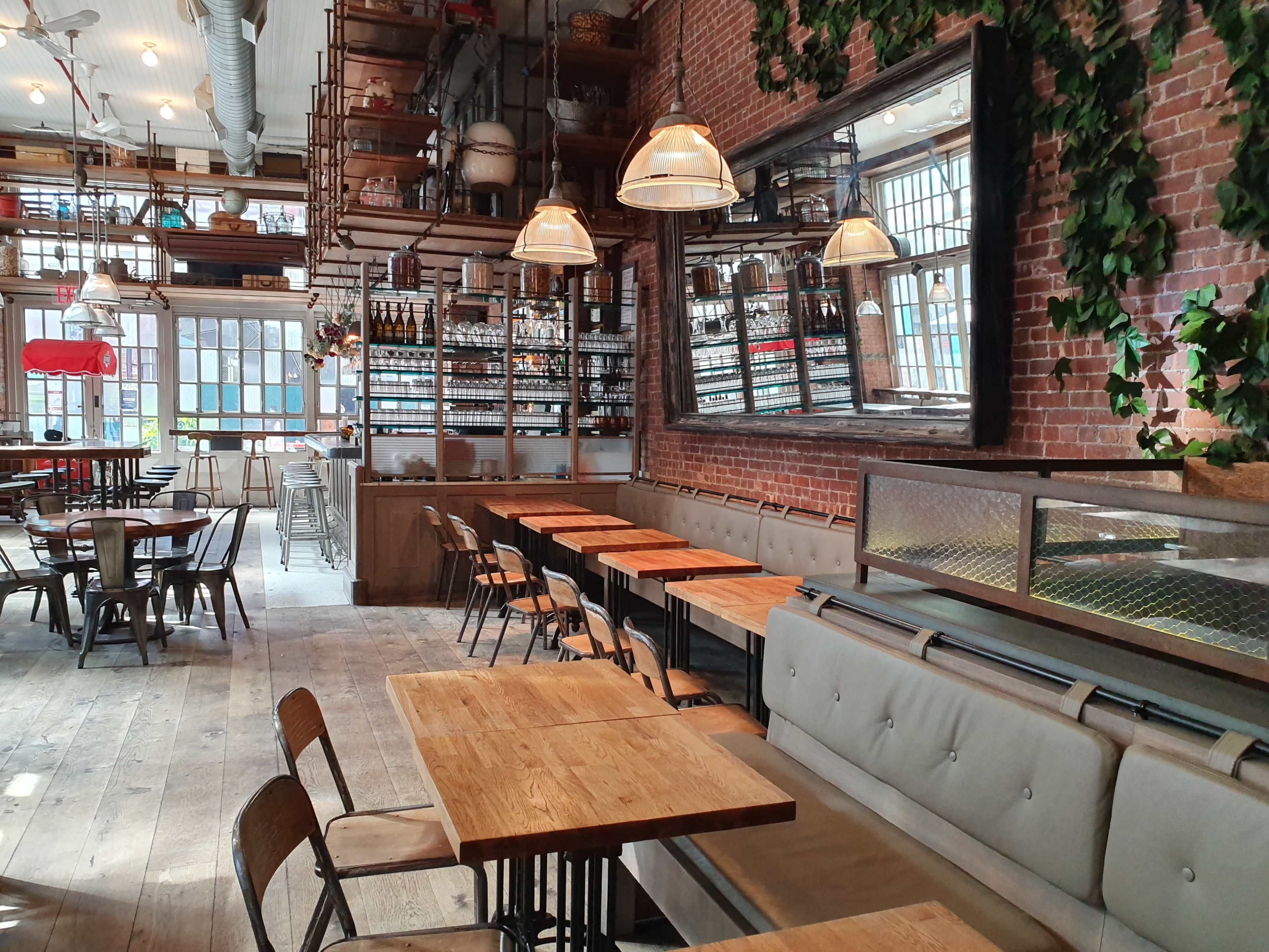 A spacious restaurant interior with wooden tables, metal chairs, and large windows, featuring a brick wall and shelves stocked with bottles.