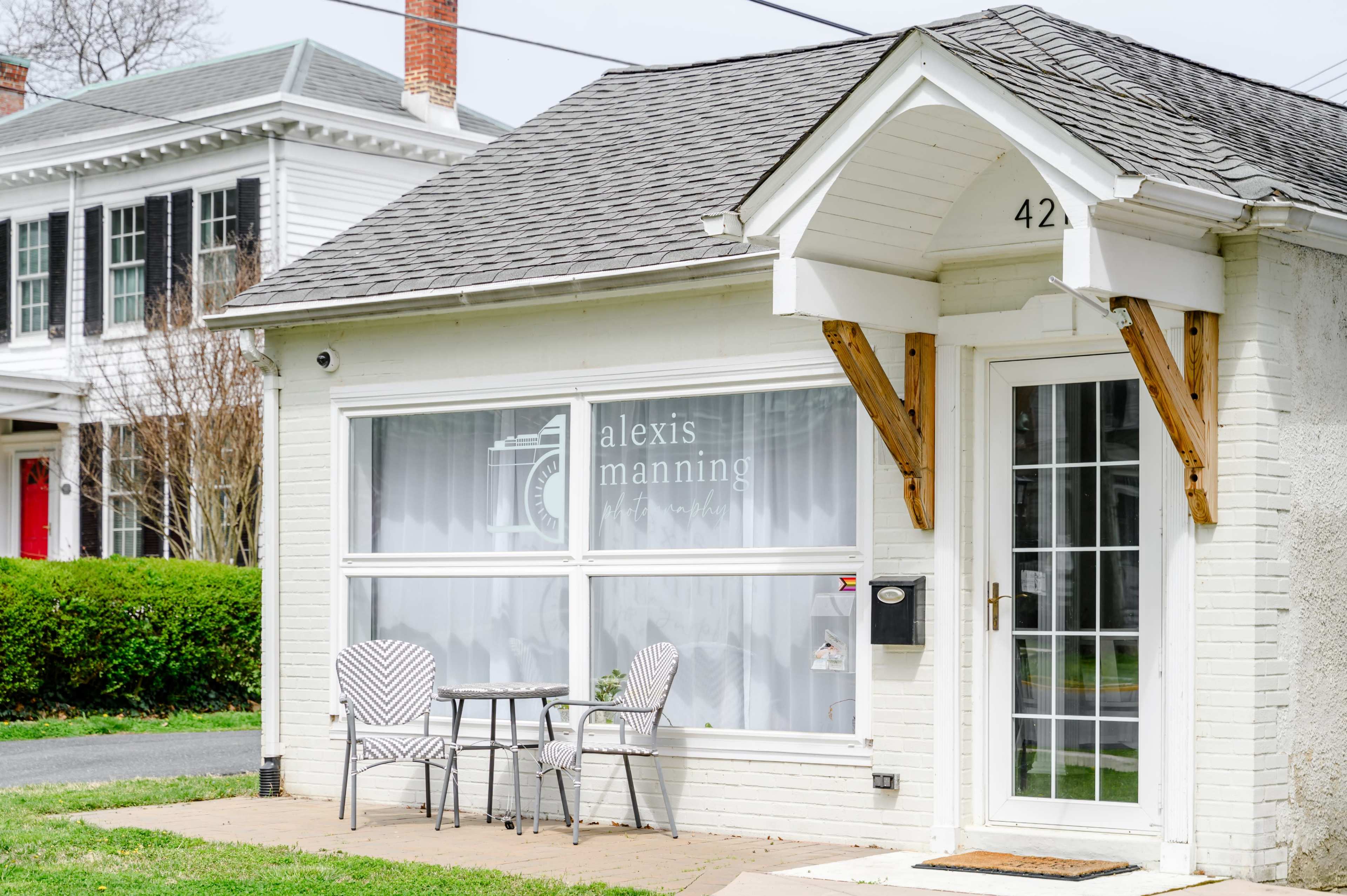 The image shows a small white building with a porch, featuring two chairs and a table, alongside a residential street.