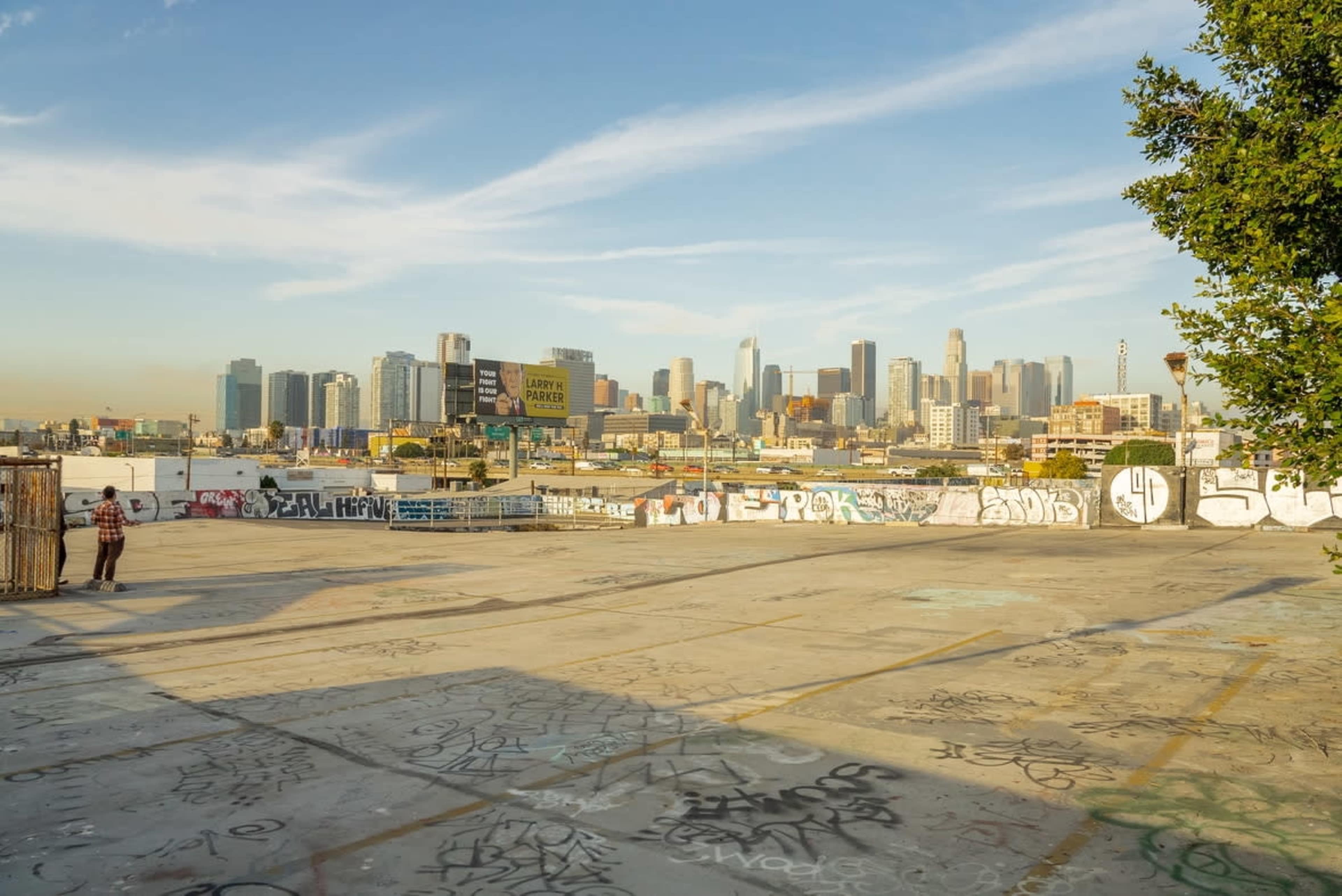 A graffiti-covered rooftop parking lot overlooks the skyline of downtown Los Angeles under a clear blue sky.