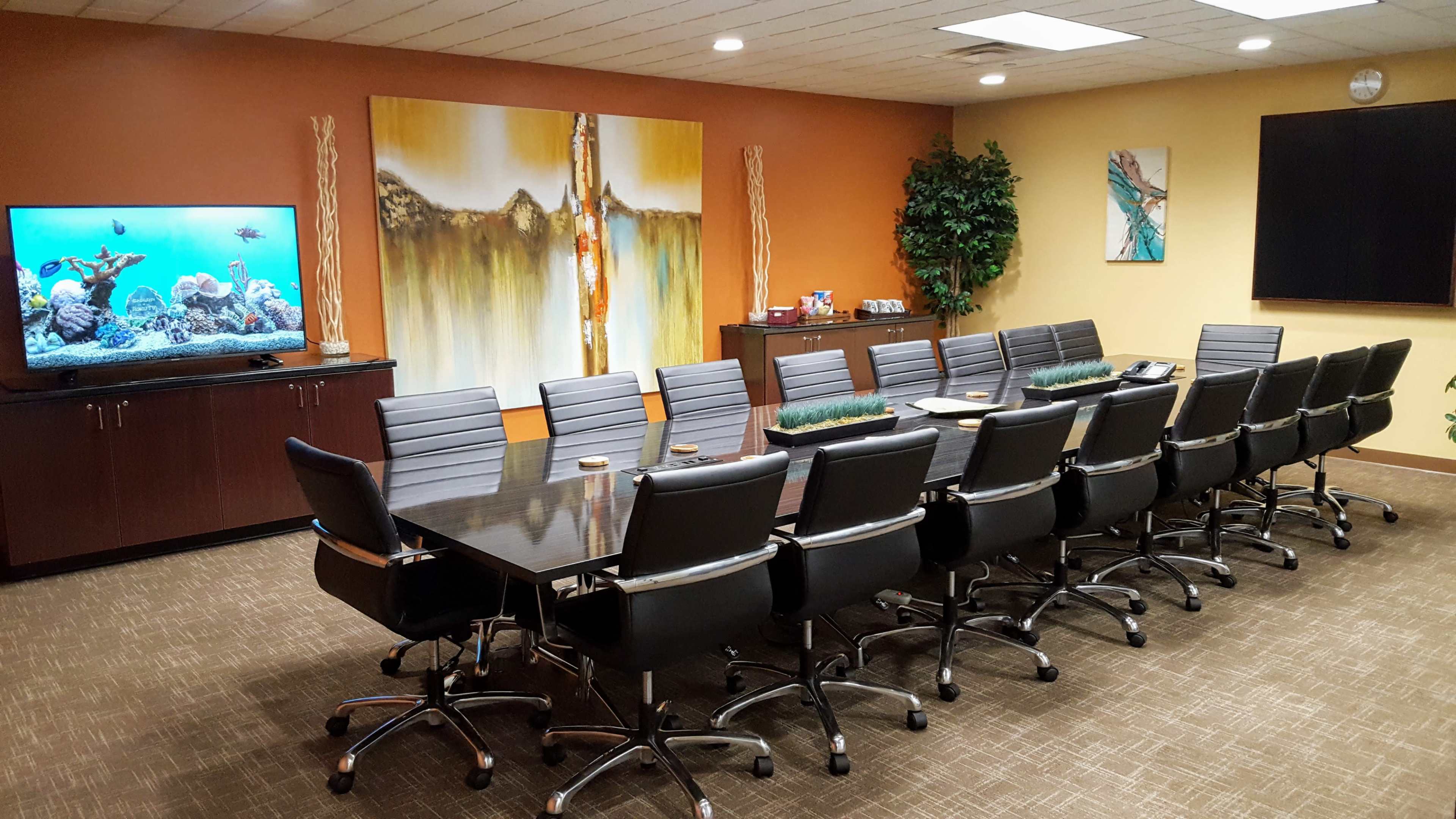 A modern conference room features a long black table surrounded by ergonomic chairs, with large artwork and a television displaying an aquarium scene on the wall.