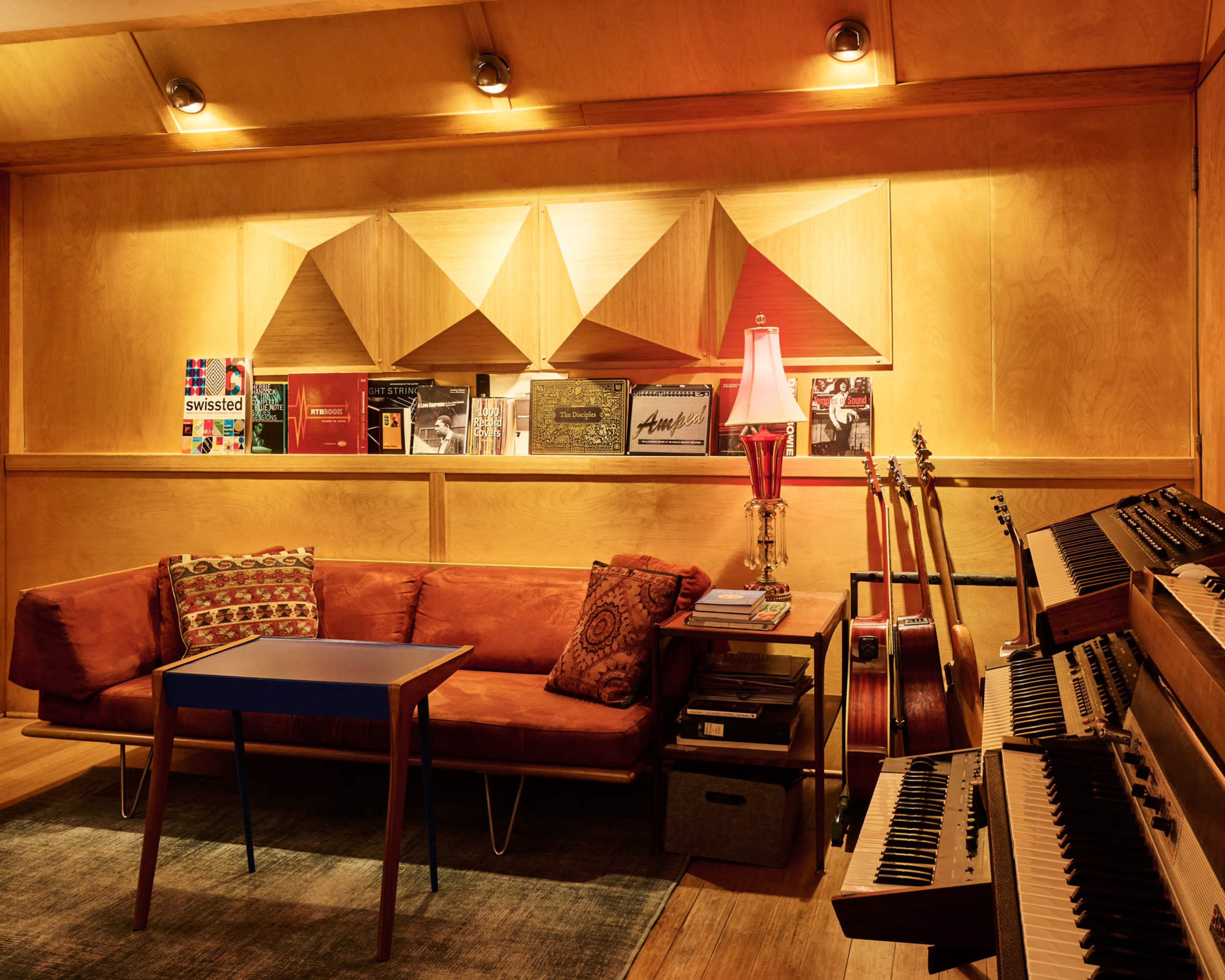 The scene features a cozy room with a brown couch, a small blue table, and a music setup, including guitars and keyboards, against a wooden backdrop with decorative acoustic panels.