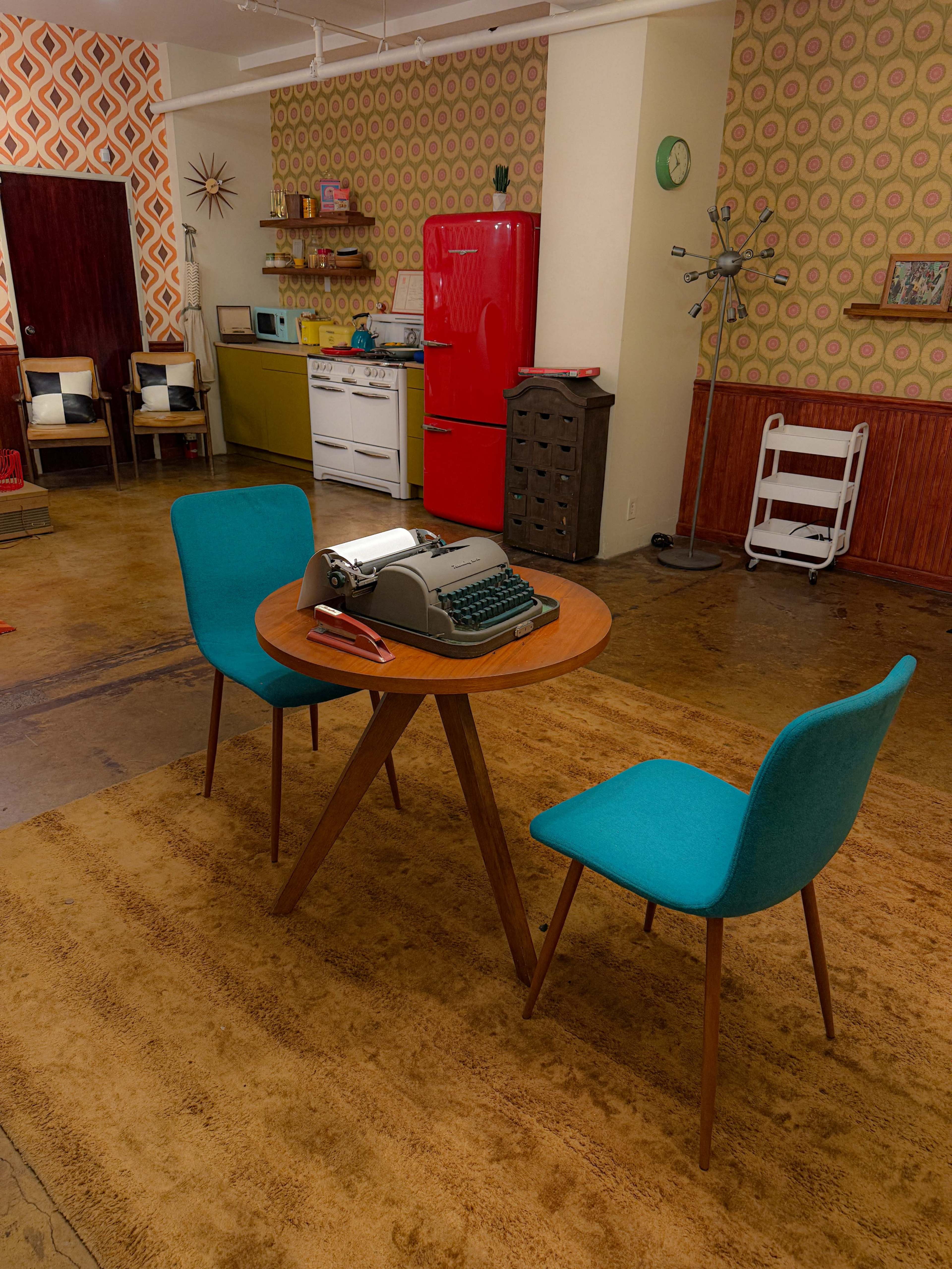 The image depicts a retro-styled room with a small wooden table, two teal chairs, and a vintage typewriter in the center.