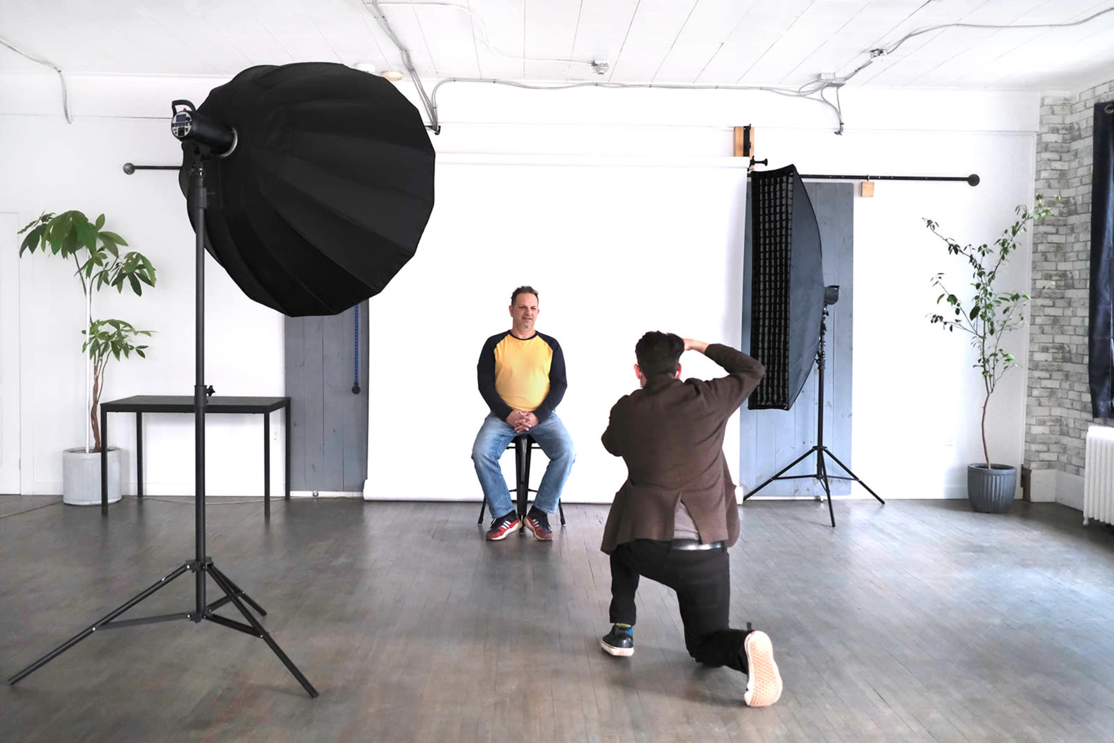 A photographer is capturing a portrait of a man sitting on a stool in a studio with a white backdrop and lighting equipment.