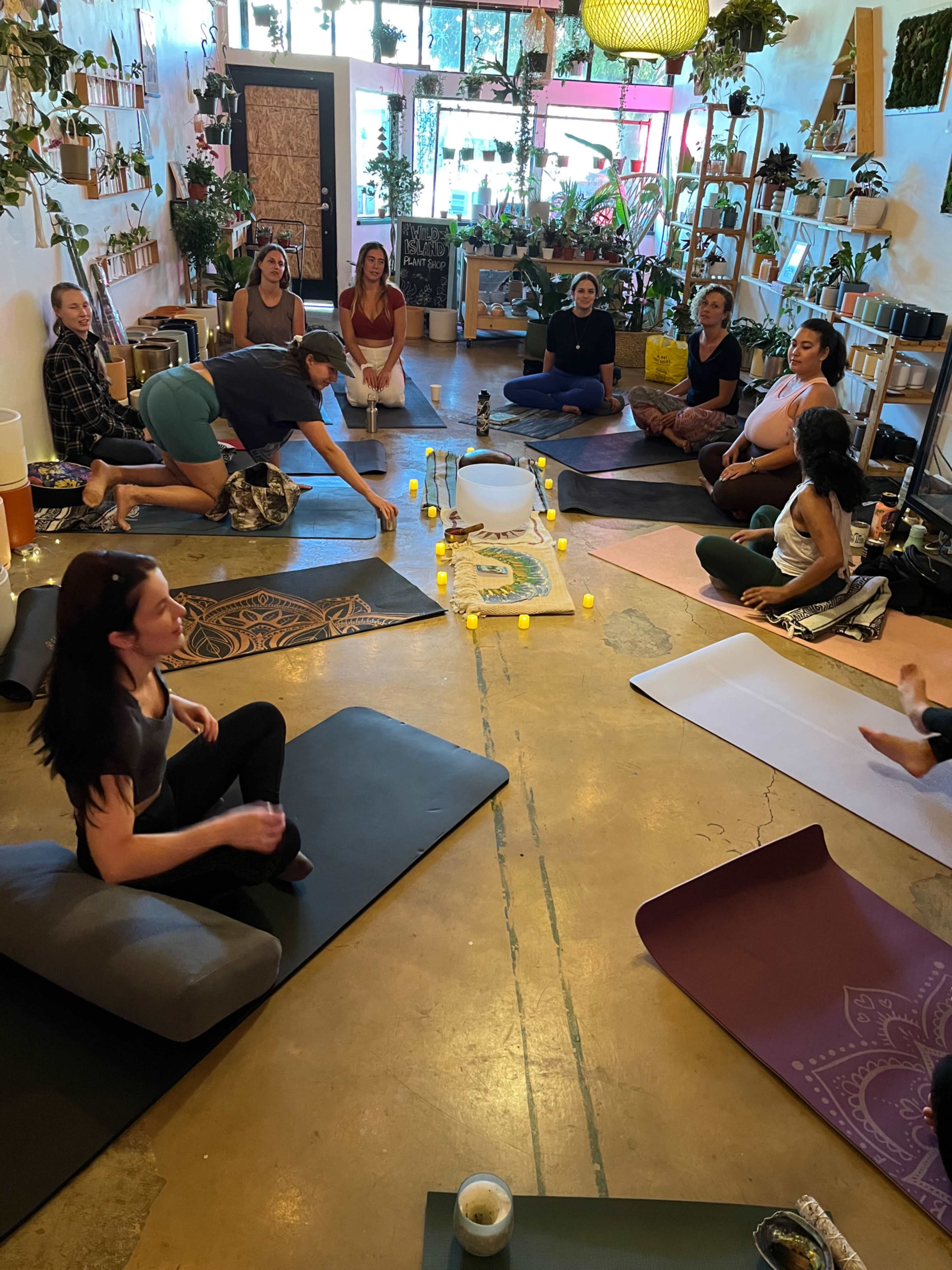 A group of individuals participates in a yoga or meditation class in a plant-filled studio, seated on mats arranged around a central layout of candles and a bowl.