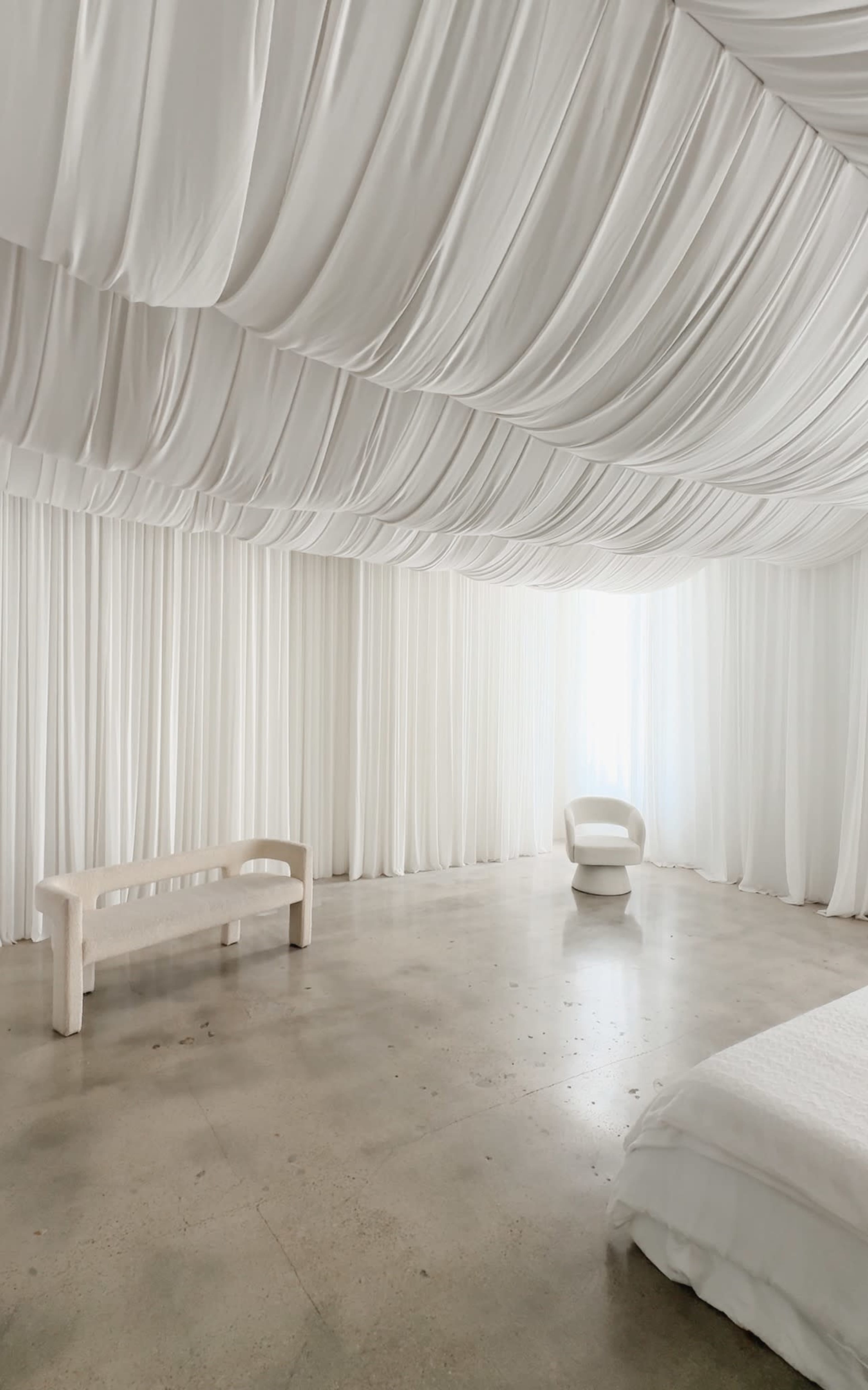 The image shows a minimalist room with draped white fabric covering the ceiling and walls, a smooth concrete floor, and simple furniture including a bench and a chair.