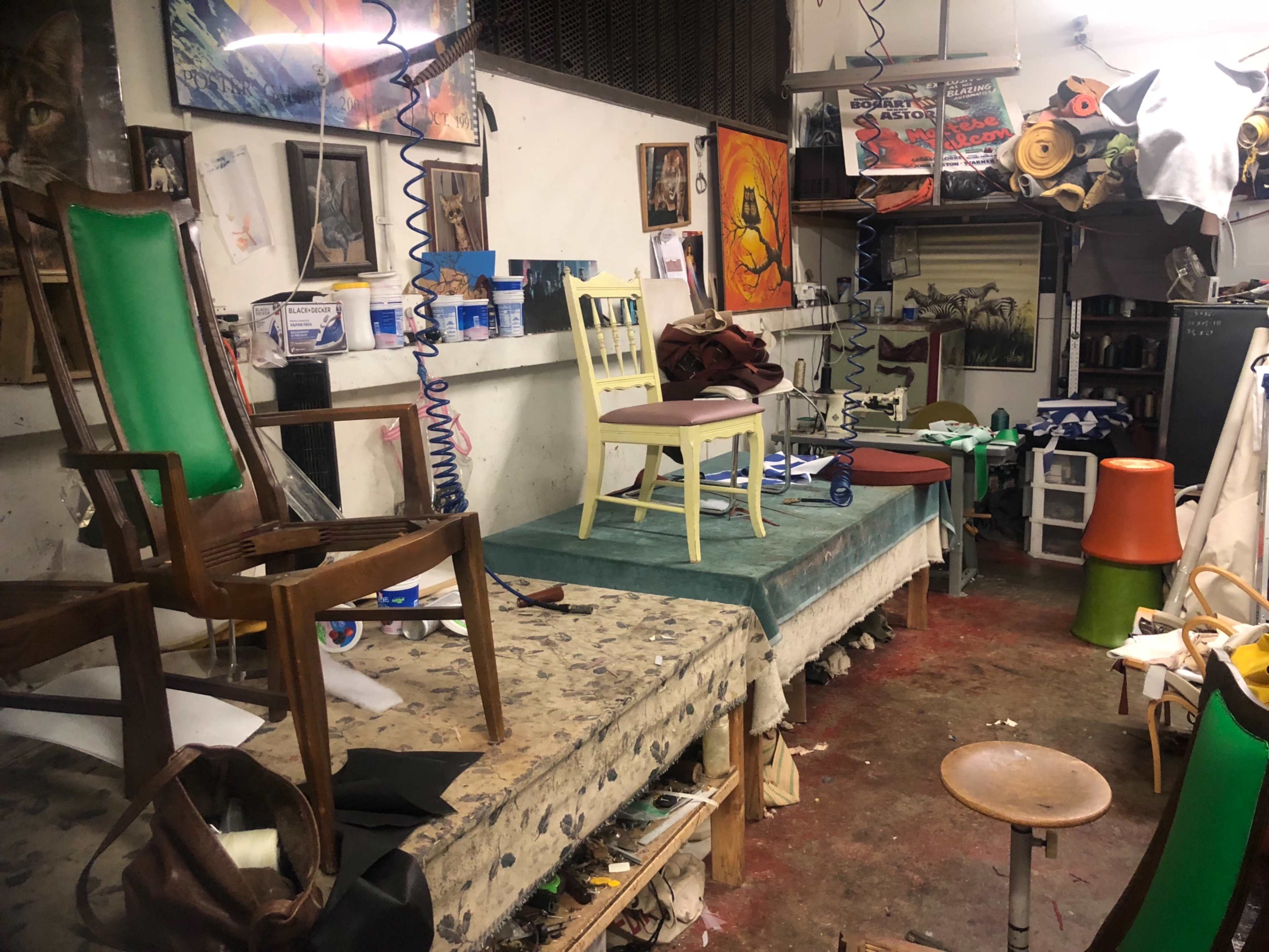 A cluttered workshop filled with various chairs, paint cans, and art supplies on tables, along with paintings on the walls.