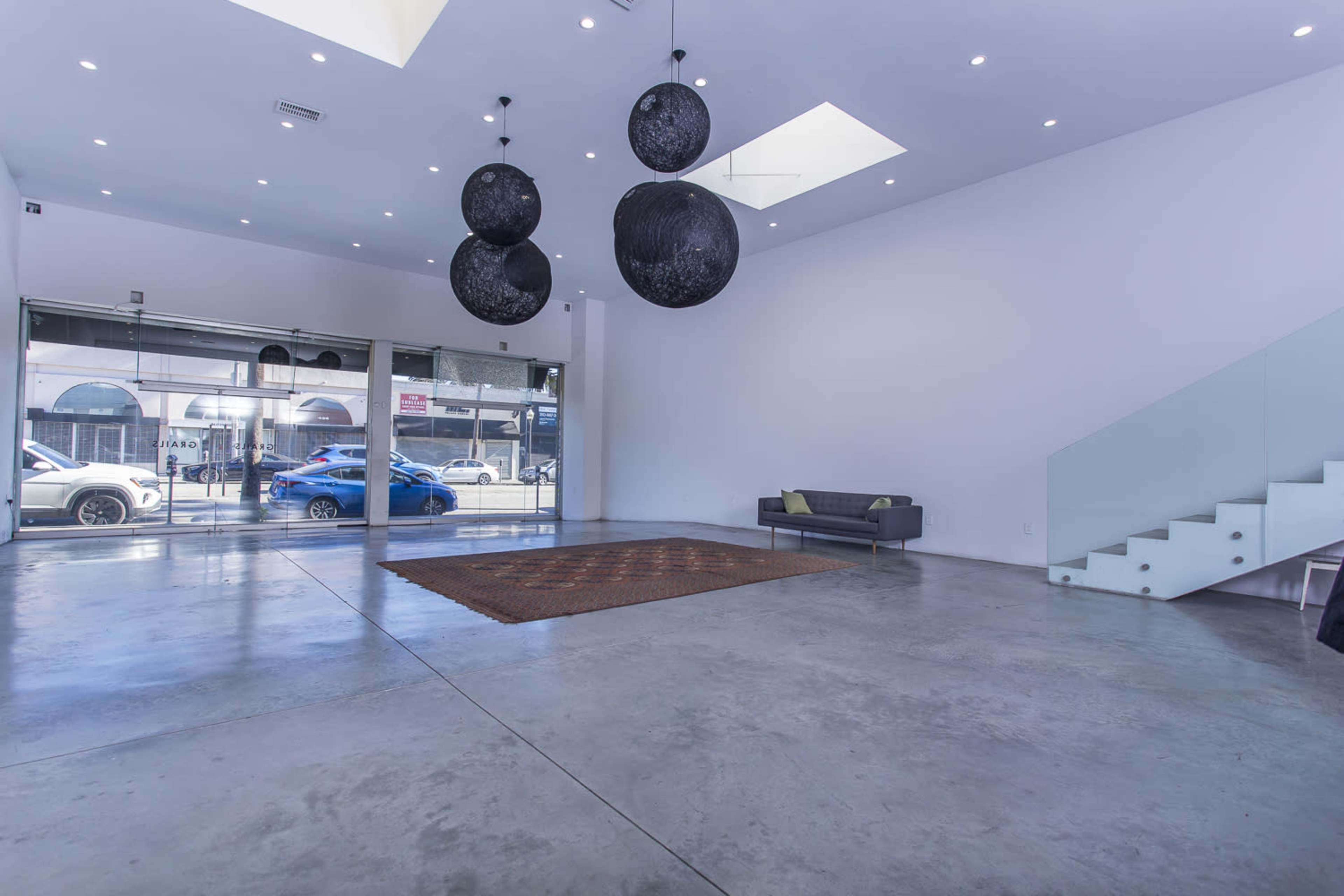The image shows a spacious, modern room with concrete flooring, large windows, and three black circular light fixtures hanging from the ceiling.