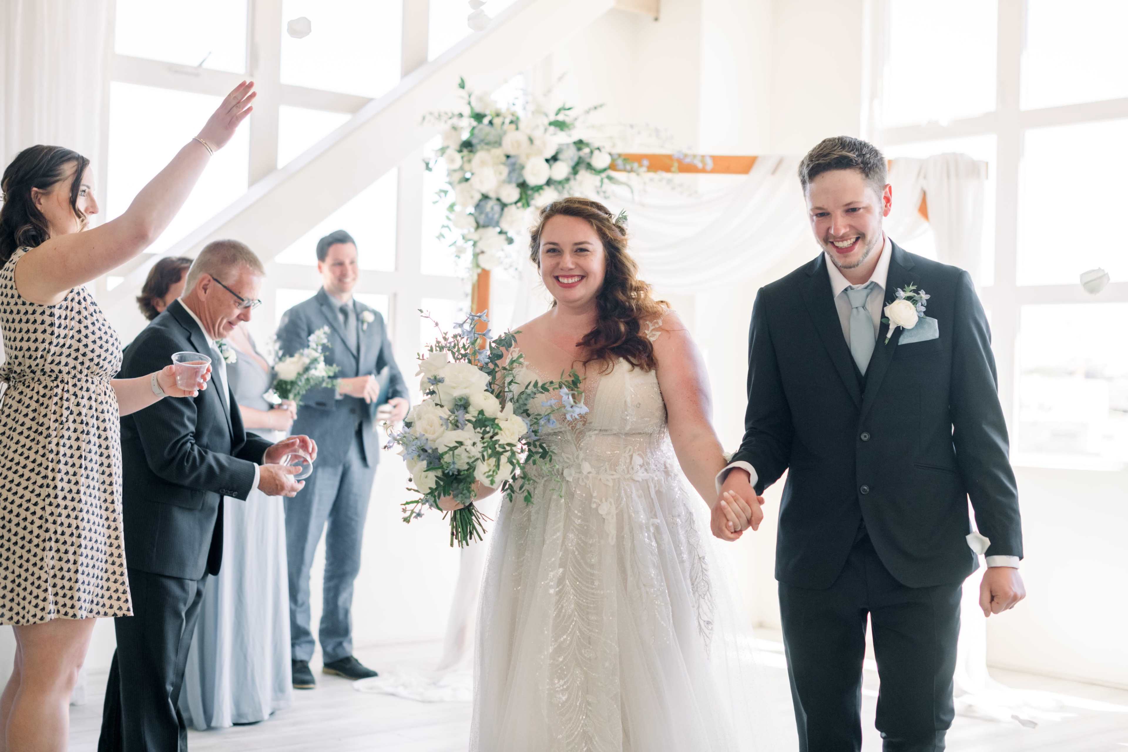 A bride and groom walk hand in hand down the aisle while guests celebrate at a wedding ceremony in a bright, airy venue.
