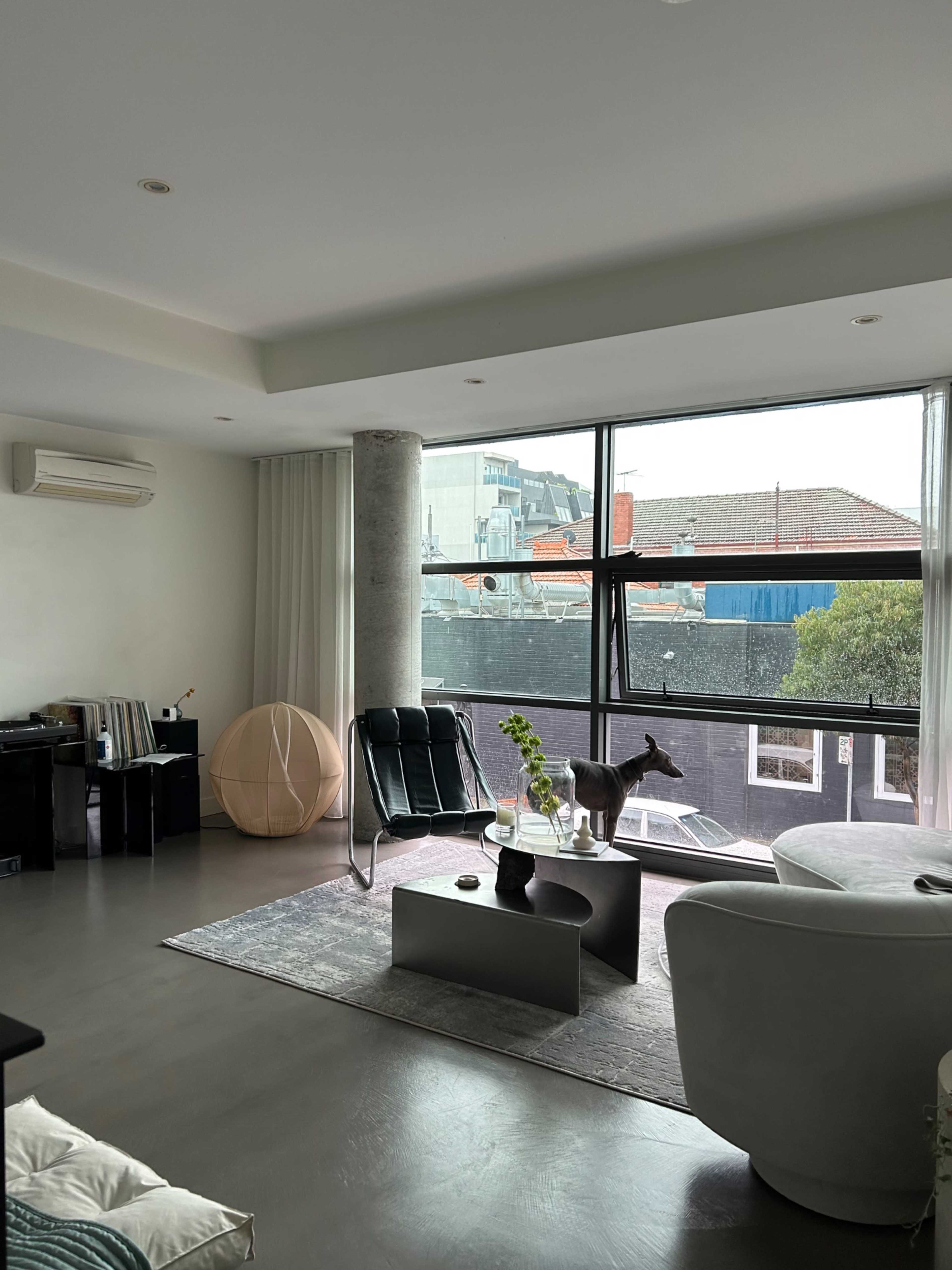 A modern living room features a large window, contemporary furniture, and a view of buildings outside.