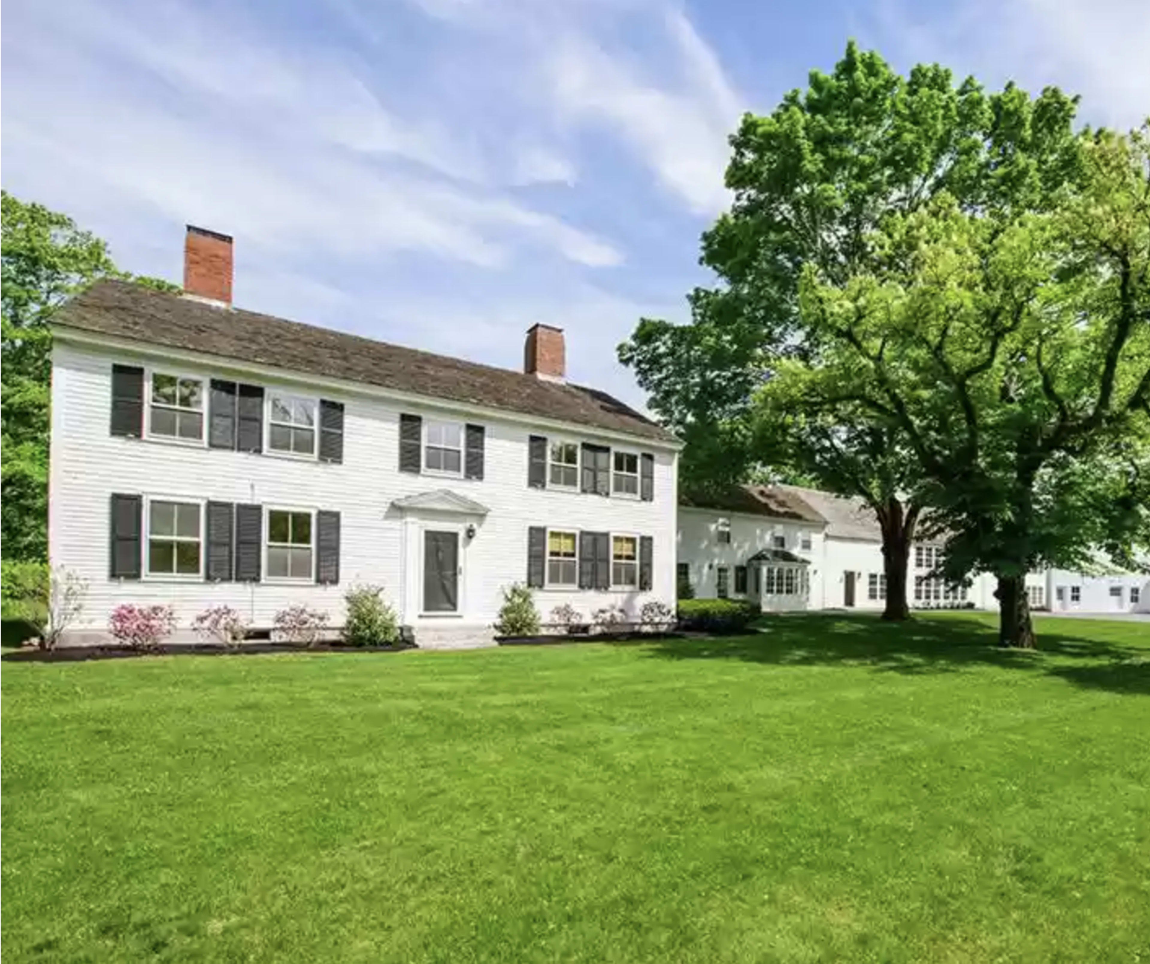 A white two-story house with black shutters surrounded by a well-maintained lawn and trees.