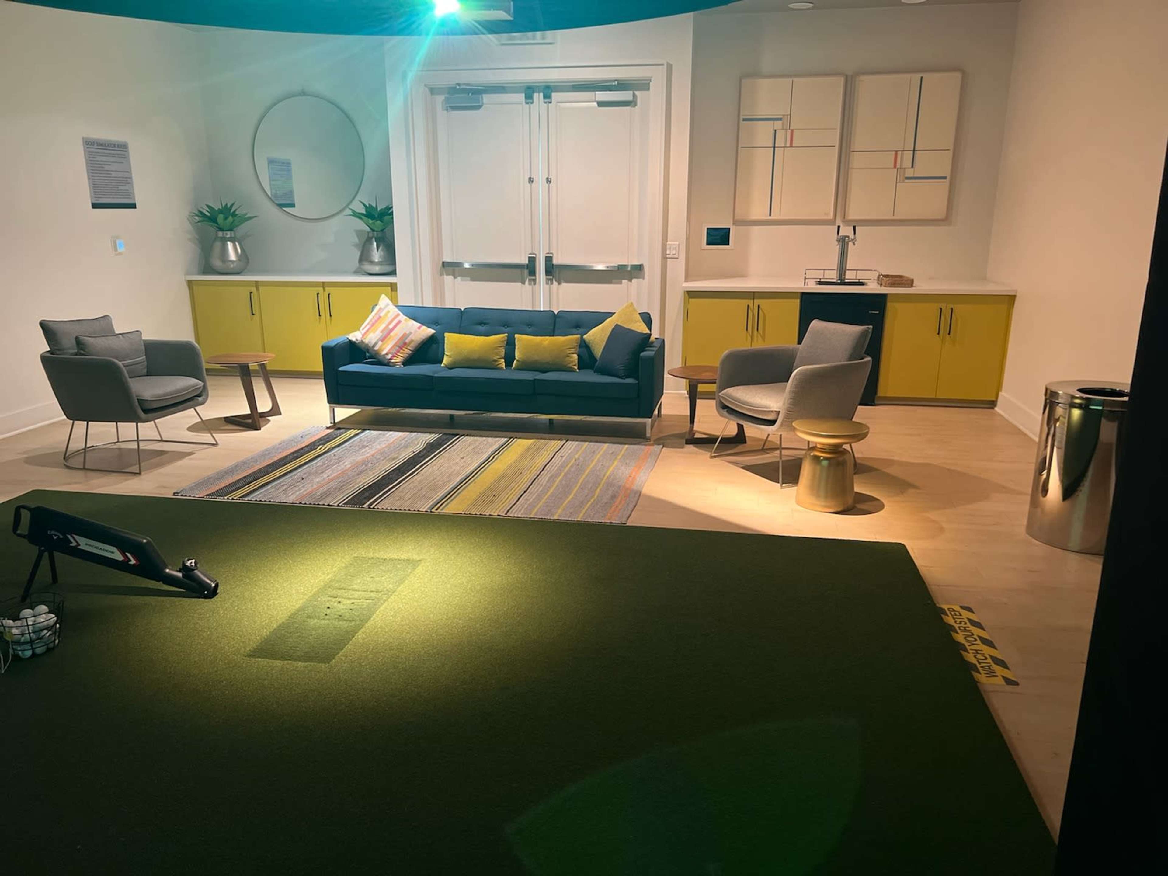 A modern indoor lounge area featuring a couch, several chairs, a small dining table, and a green putting surface with golf equipment.