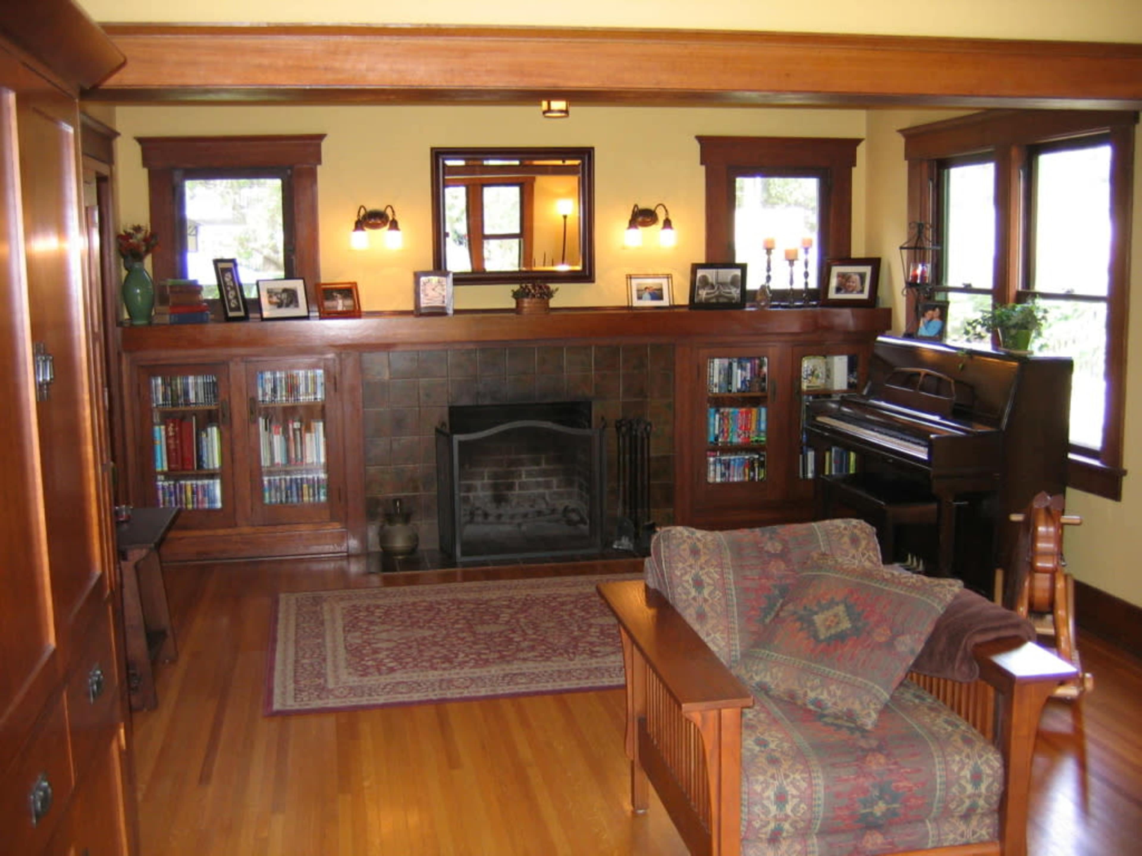A cozy living room with wooden beams, a fireplace, a piano, bookshelves filled with books, and a patterned rug.