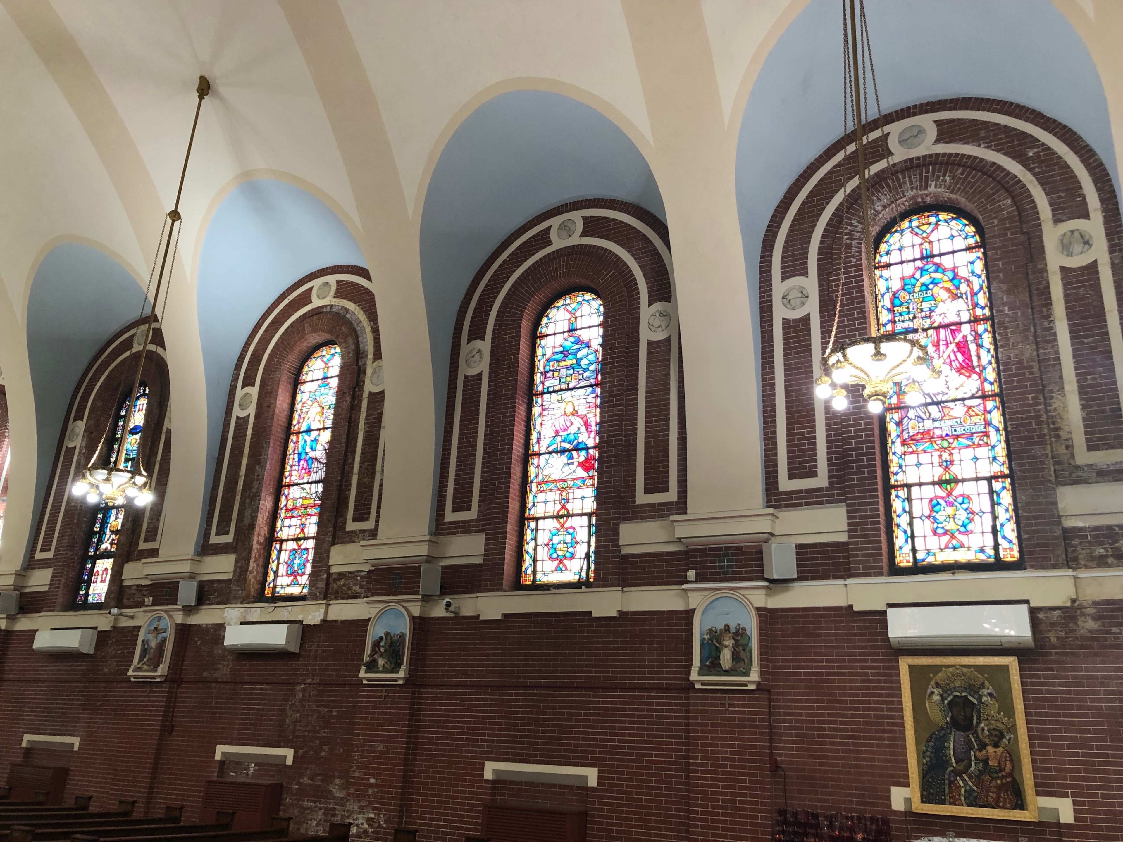 The interior of a church, featuring arched stained glass windows and decorative lighting fixtures along the walls.