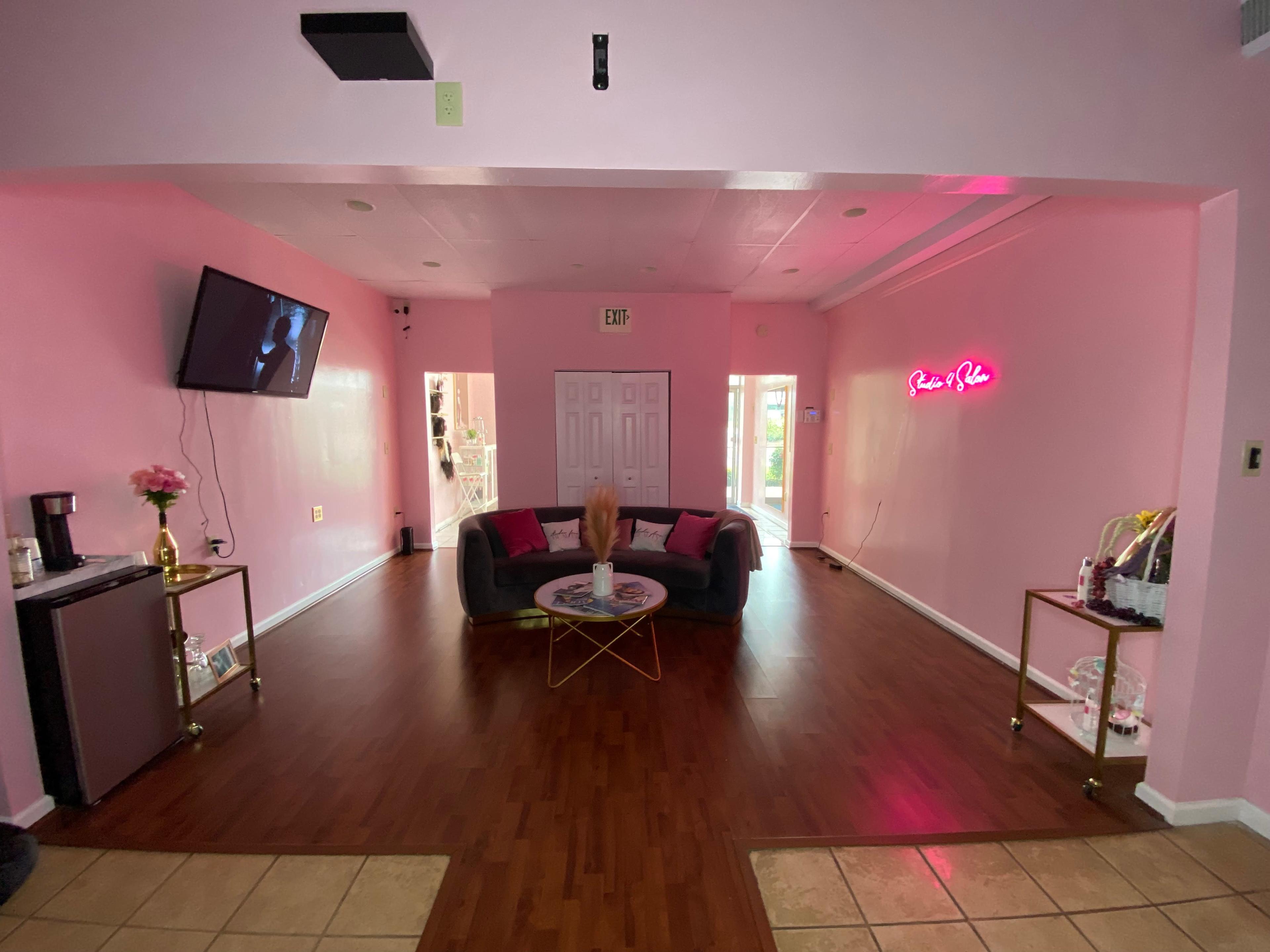 A pink-walled room with a couch, a television mounted on the wall, and a neon sign, featuring a coffee station and a decorative table.
