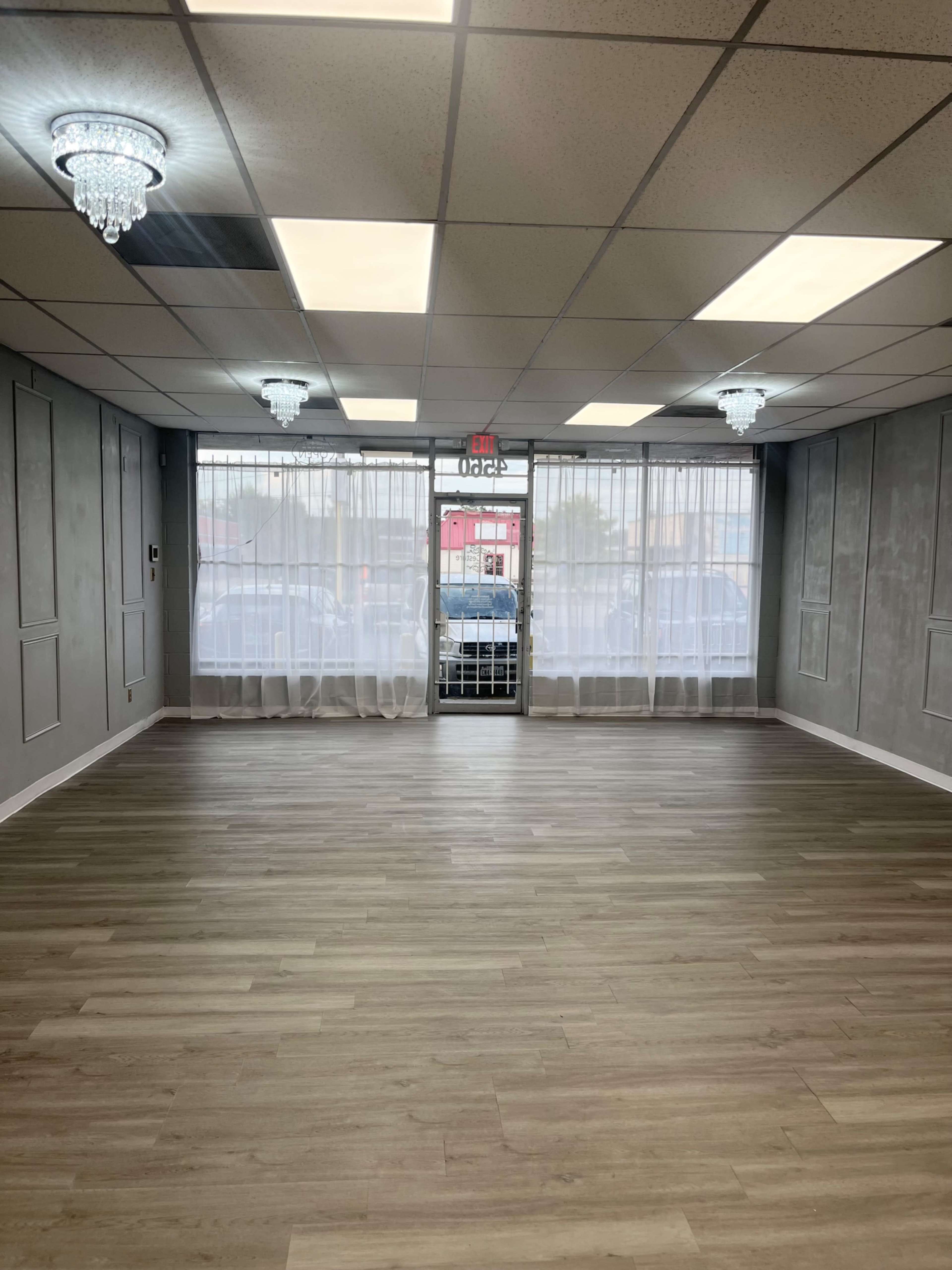 The image shows an empty interior room with hardwood flooring, light gray walls, large windows covered with sheer curtains, and two crystal light fixtures on the ceiling.